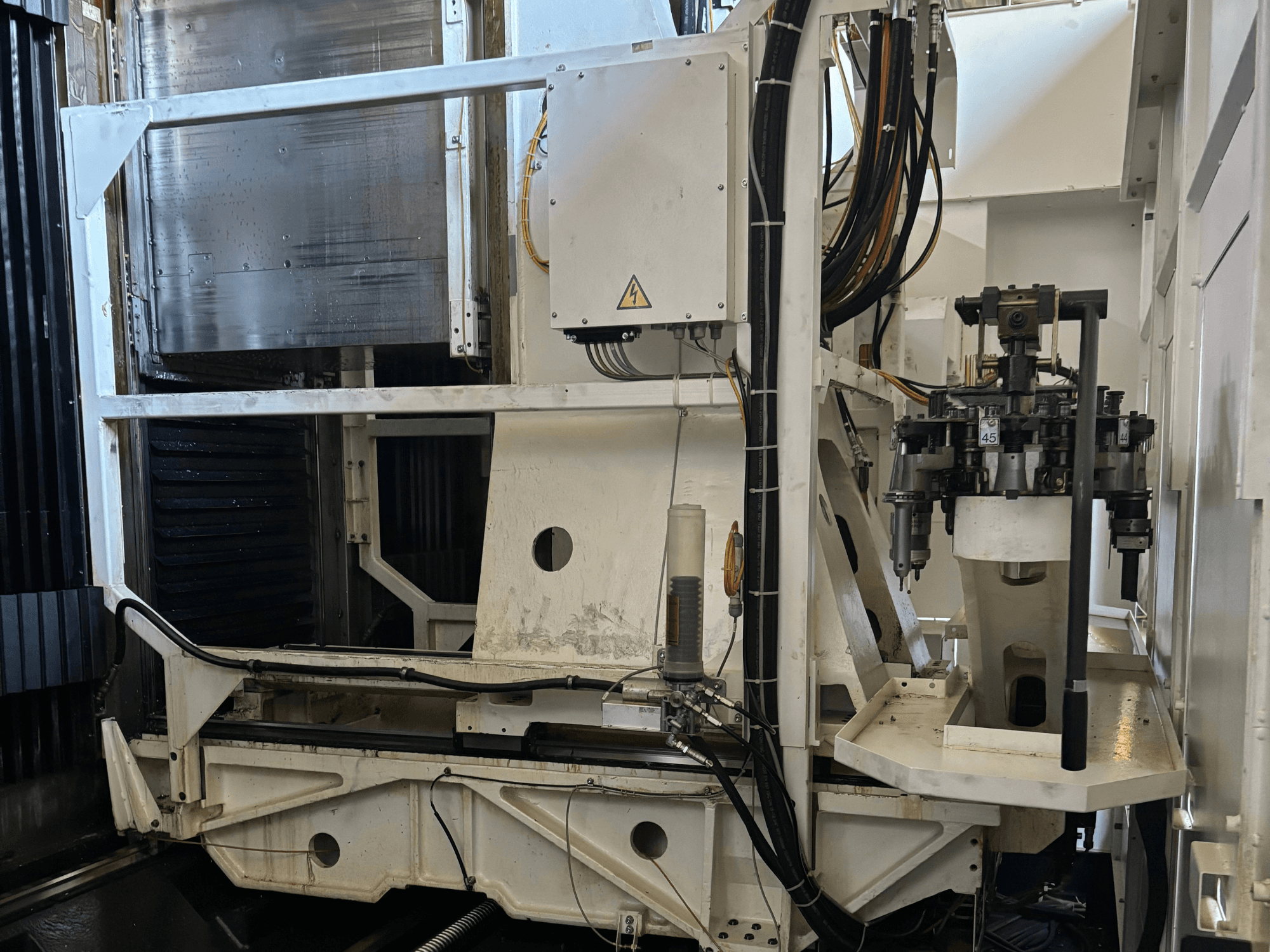 Ipari gép oldalnézetben egy fehér CNC marógép látható szerszámokkal, csövekkel és vezérlőpanellel.