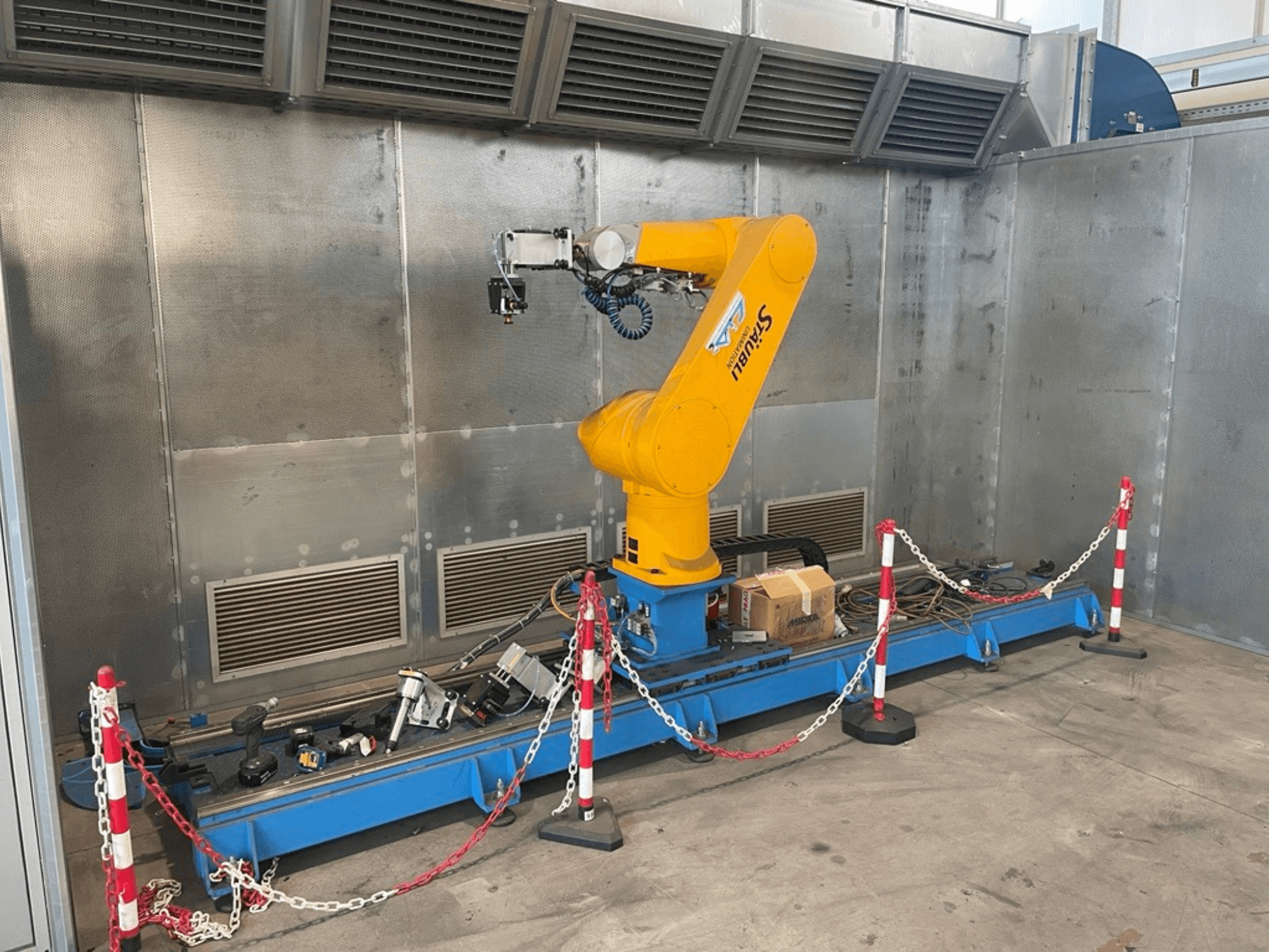 STAUBLI CS8C-RX160 robotkar műhelyben, oldalnézetben, kábelekkel és különböző szerszámokkal, kék alapon, védőfalakkal körülvéve.