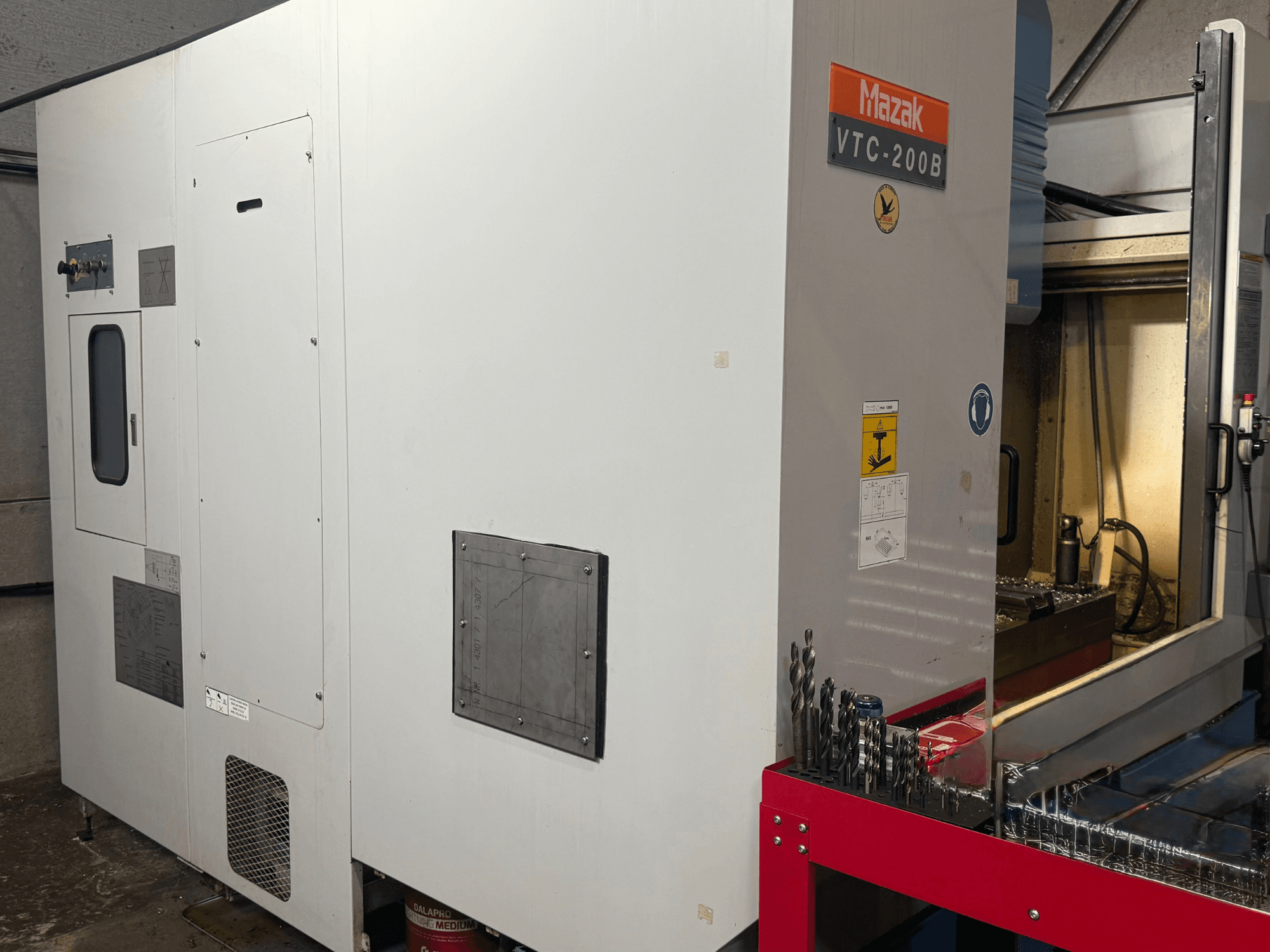 A Mazak VTC-200B függőleges megmunkálóközpont oldalnézete, feliratozott kezelőszervekkel és a piros tárolóállványon elhelyezett fúrófejekkel.