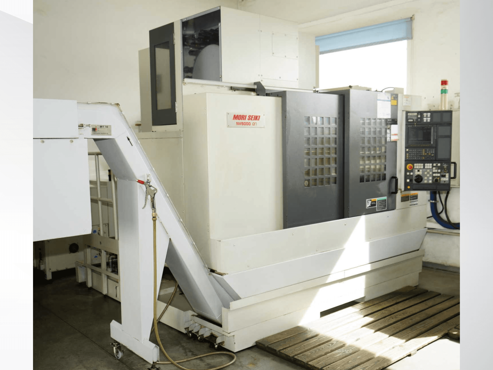 A MORI SEIKI NV 5000 alpha1 függőleges megmunkáló központ elölnézete, amelyen látható a vezérlőpanel, a biztonsági berendezések és a betöltőterület.