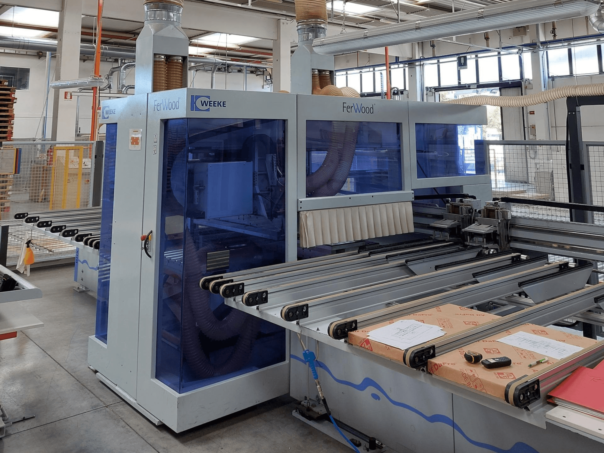 Weeke FerWood CNC-gép egy műhelyben, elölnézetben a kék színű panelek, a vezérlőgombok és a munkaasztal az anyagokkal.