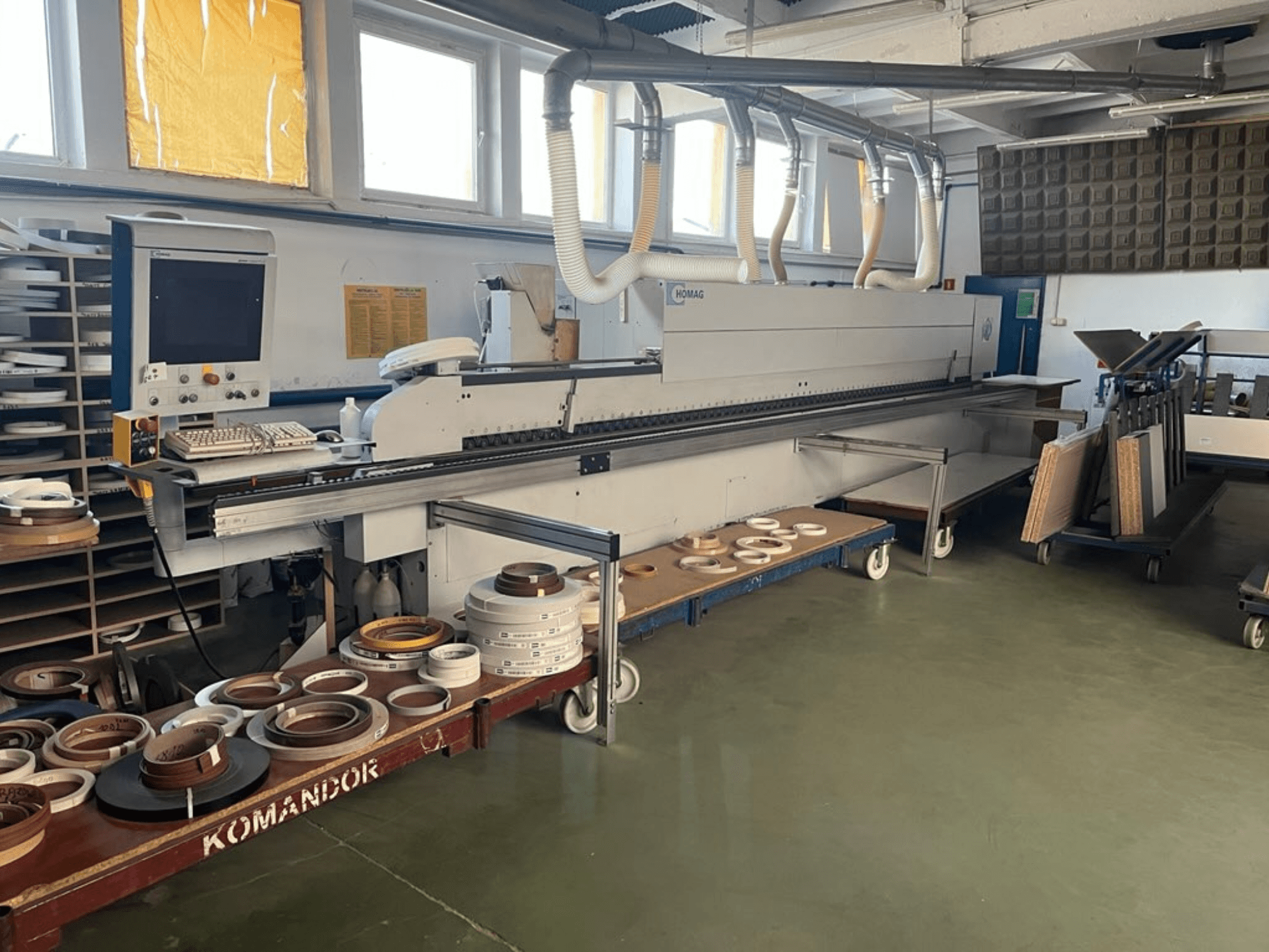 HOMAG élszalagozó gép egy műhelyben, kezelőszervekkel és különböző élszalagozó anyagokkal egy kék kocsin. Oldalnézet.