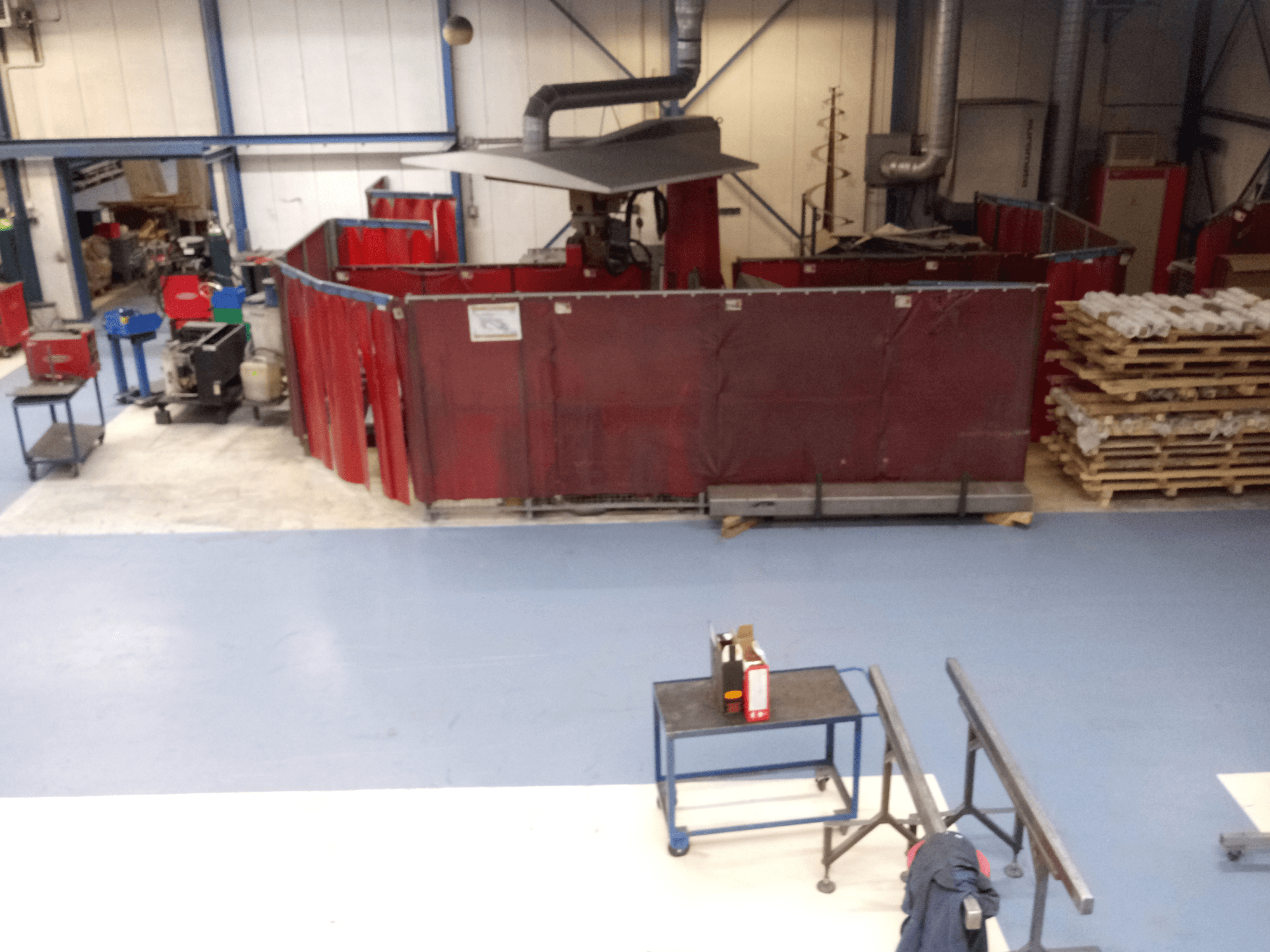 Ipari munkaterület piros hegesztőfüggönyökkel, gépekkel és raklapokkal; felülnézetben az elrendezés és a berendezések bemutatása.