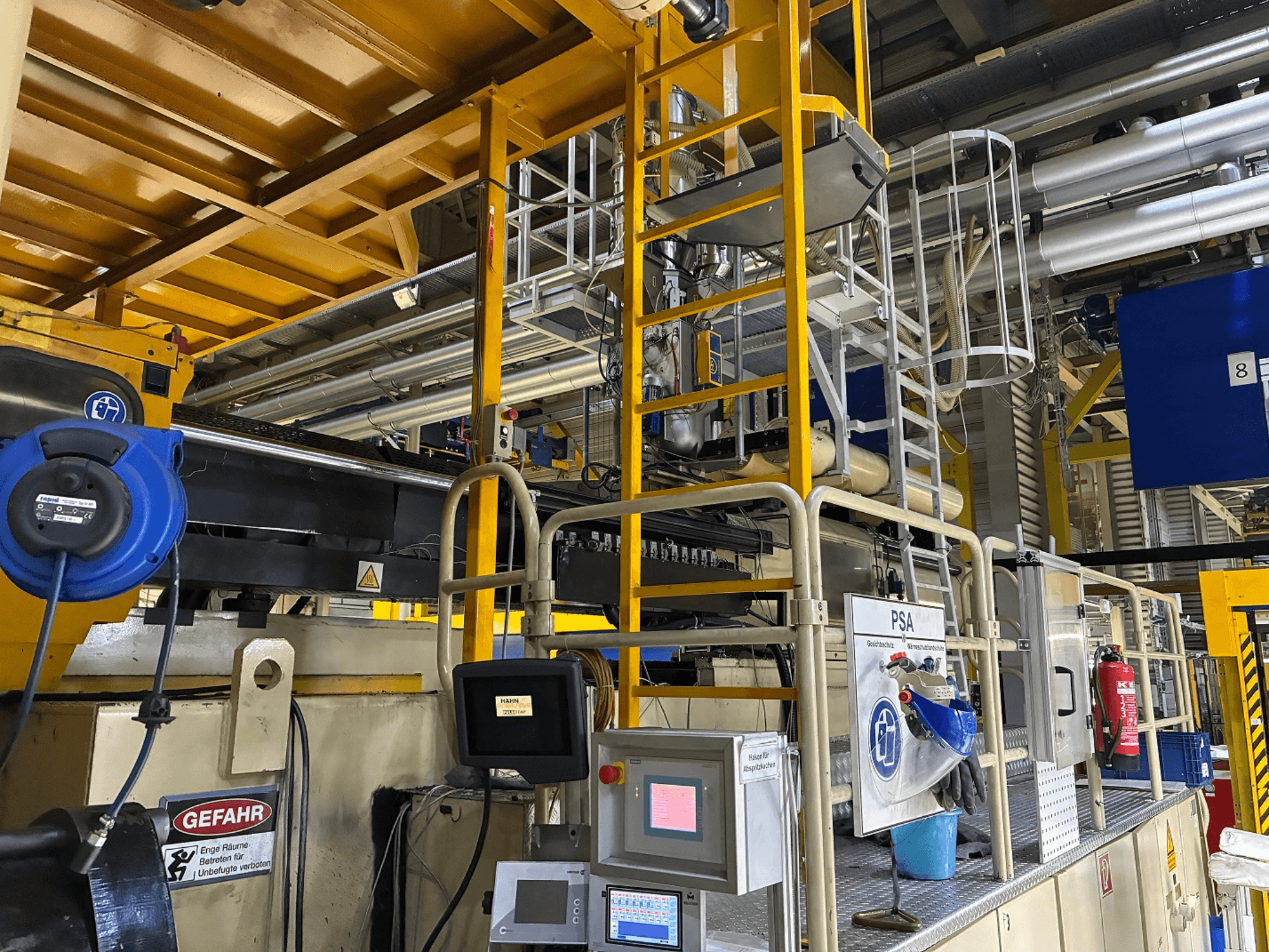 Ipari gépek vezérlőpultja kék biztonsági felszereléssel és sárga állványzattal, oldalnézetben csövek és figyelmeztető táblák.