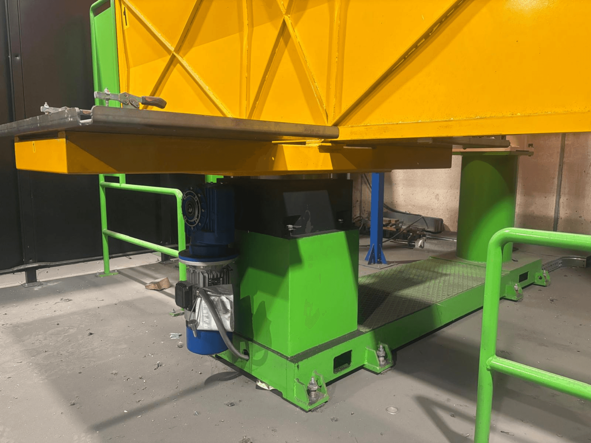 Zöld és sárga színű ipari gép alacsony elülső nézetből, motorral és texturált felületű platformmal.