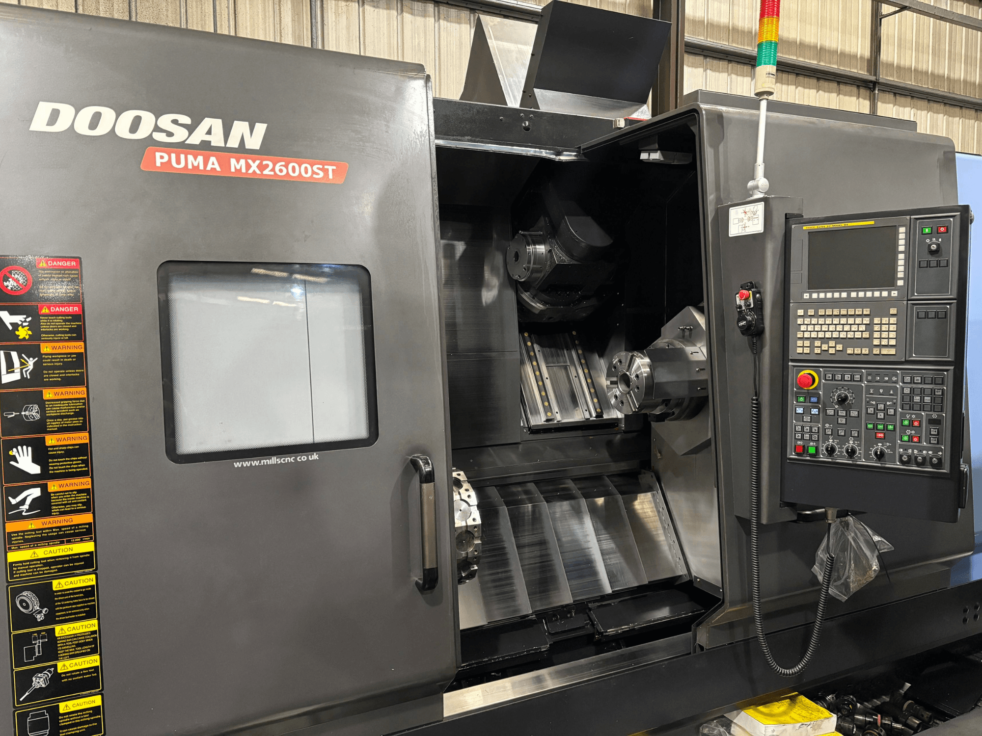 Doosan PUMA MX2600ST CNC eszterga, előlnézet, a vezérlőpanel, az orsó és a megmunkálási terület látható biztonsági figyelmeztetésekkel.