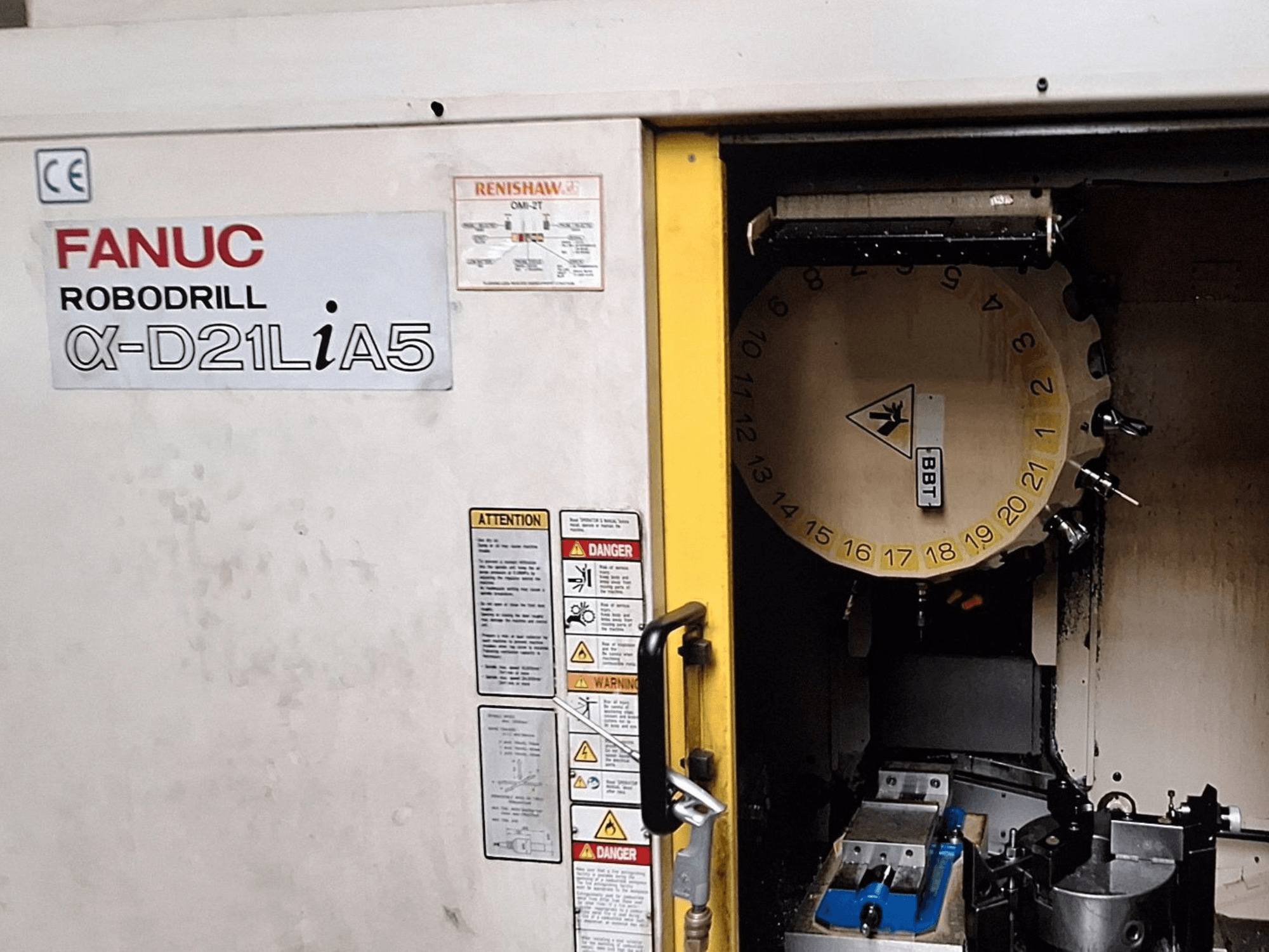 FANUC Robodrill α-D21LiA5, előlnézet. A szerszámkarusszel és a megmunkálási területet mutatja a rögzítőkkel és az ajtón lévő figyelmeztetésekkel.