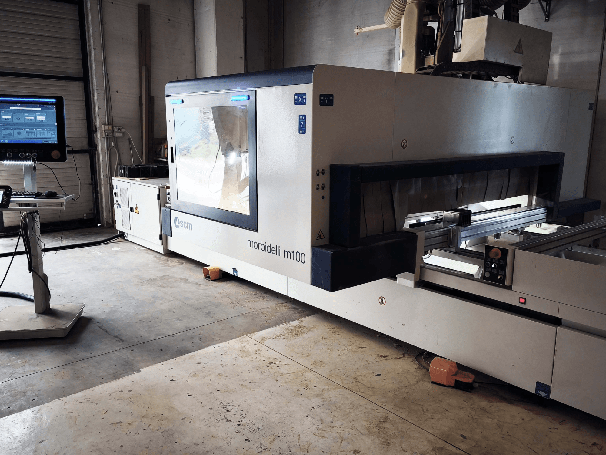 Az SCM Morbidelli M 100 CNC famegmunkáló központ oldalnézete, amelyen látható a vezérlőpanel, a munkaterület és a biztonsági berendezések.