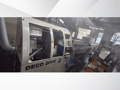 DECO 2000 CNC-gép egy műhelyben, elölnézetben, a kezelőszervek és egy hosszú munkadarab beállítása a jobb oldalon.