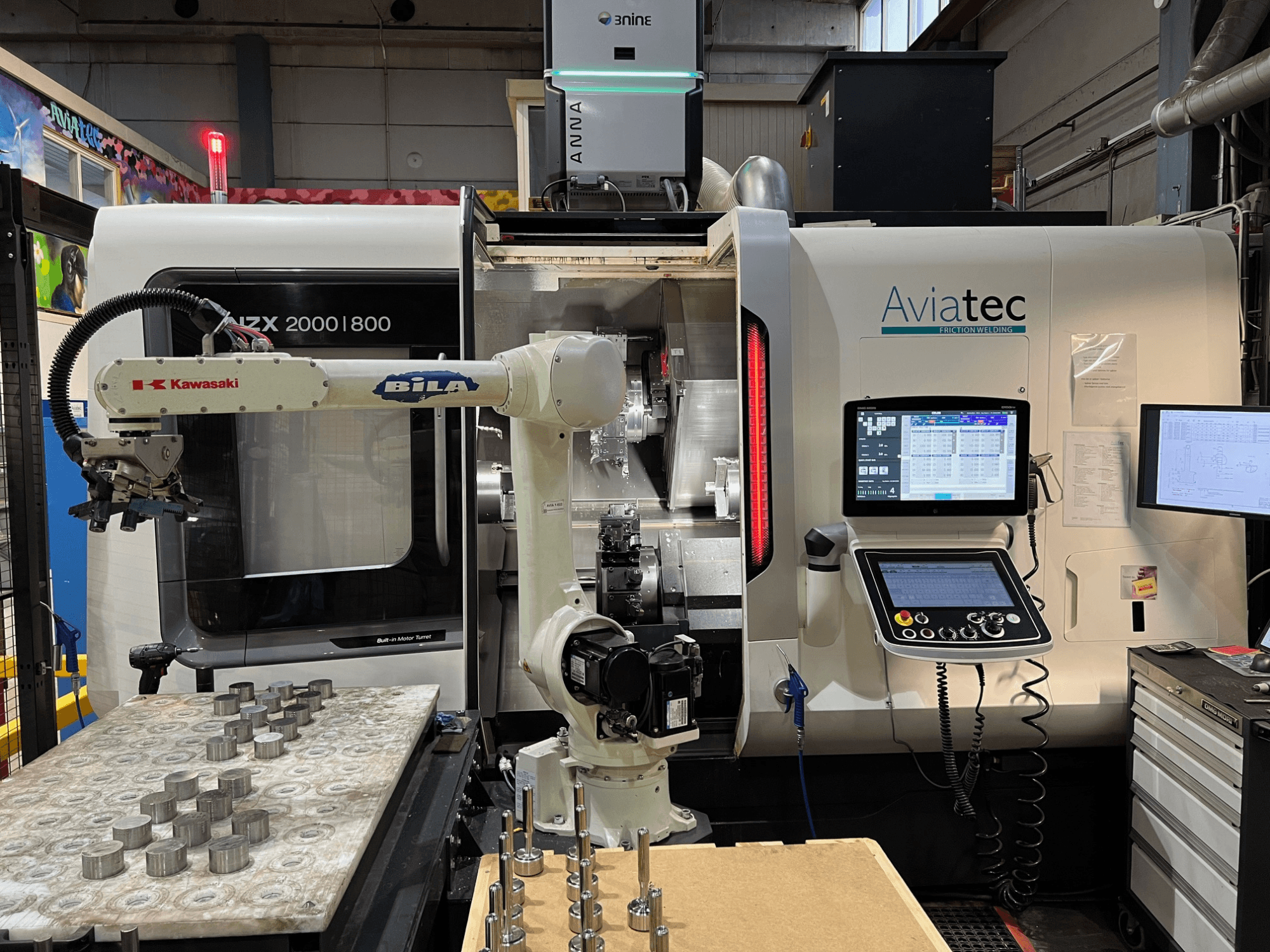 DMG MORI NZX 2000/800SY vízszintes eszterga Kawasaki robotkarral, vezérlőpanellel és a munkapadon elhelyezett fém alkatrészekkel.