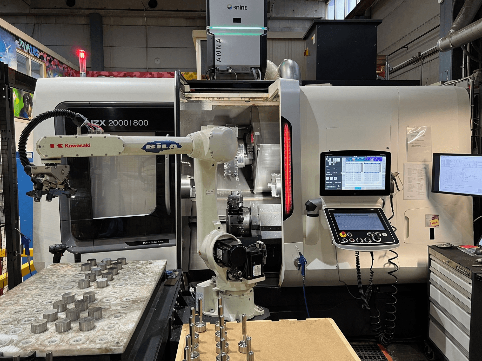 DMG MORI NZX 2000/800SY vízszintes eszterga egy műhelyben, látható a Kawasaki robotkar és a vezérlőegységek.