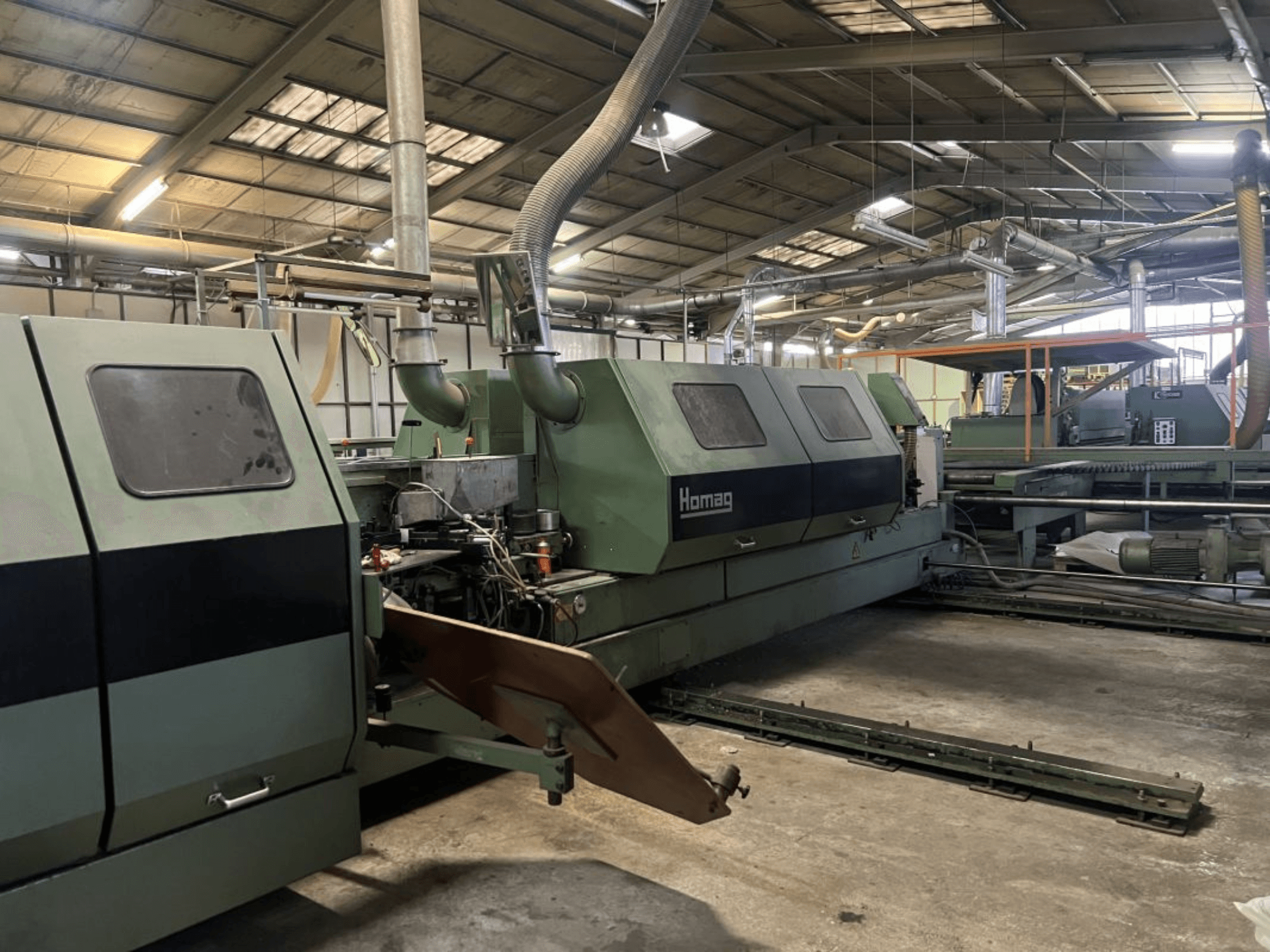 Homag ipari gép zöld színben, oldalnézetben, láthatóak a működő alkatrészek és a vezérlőpanel egy raktárban.