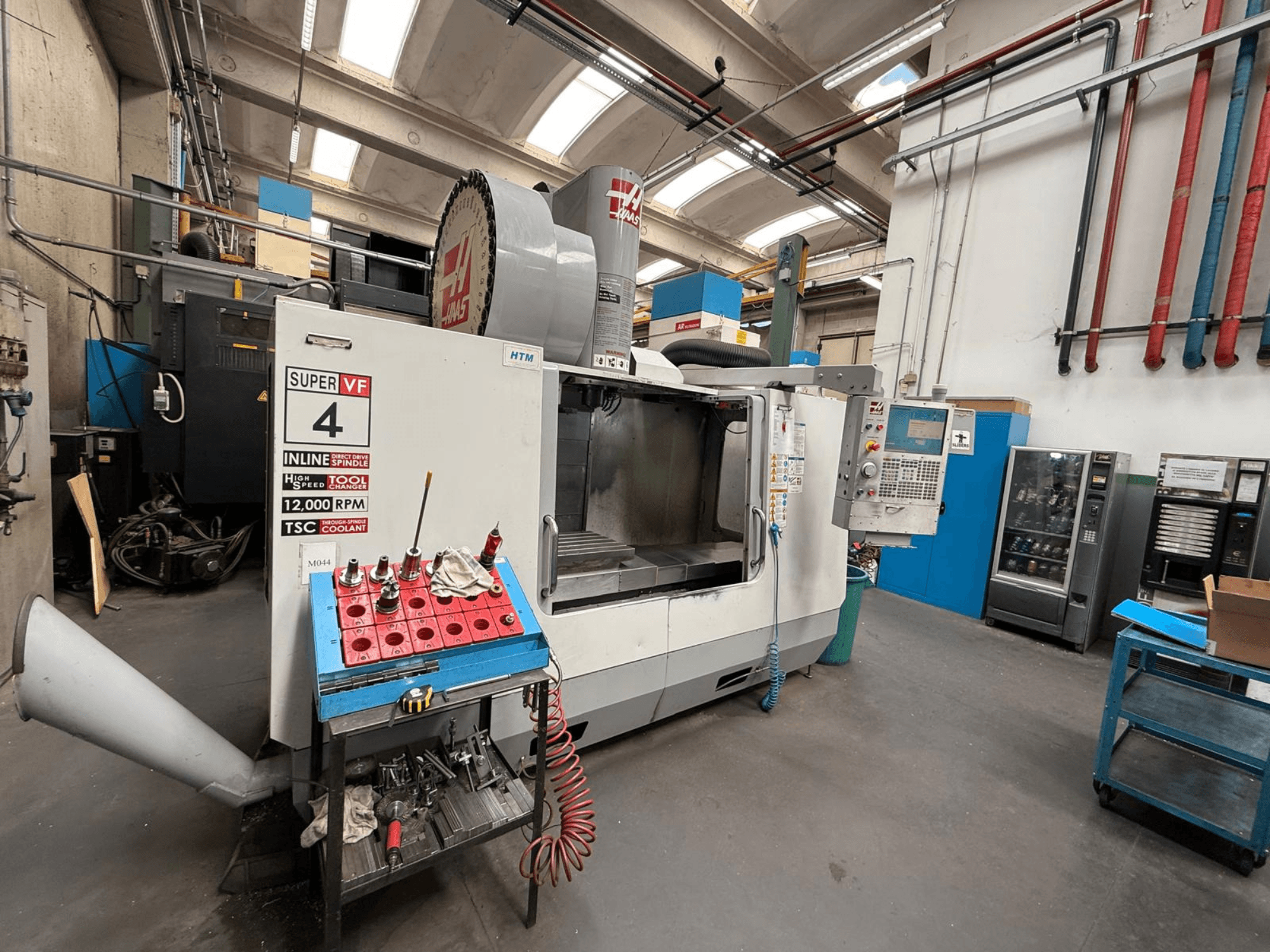 HAAS Super VF 4 CNC marógép vezérlőpanellel, 12 000 fordulat/perc fordulatszámú orsóval és szerszámokkal egy kék tárolóállványon, oldalnézet.