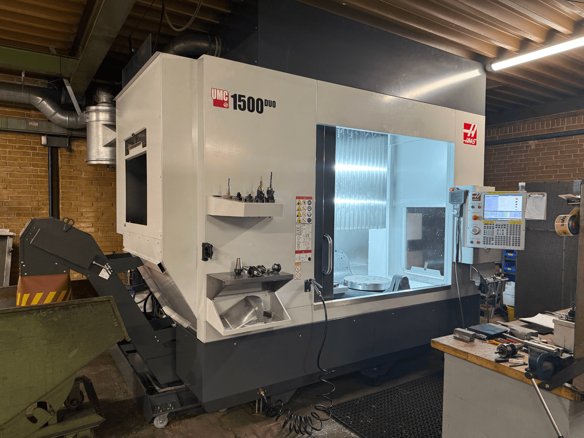 Haas UMC 1500DUO CNC-gép, előlnézet, nagy ablak, vezérlőpanel és szerszámtartók egy polcon.
