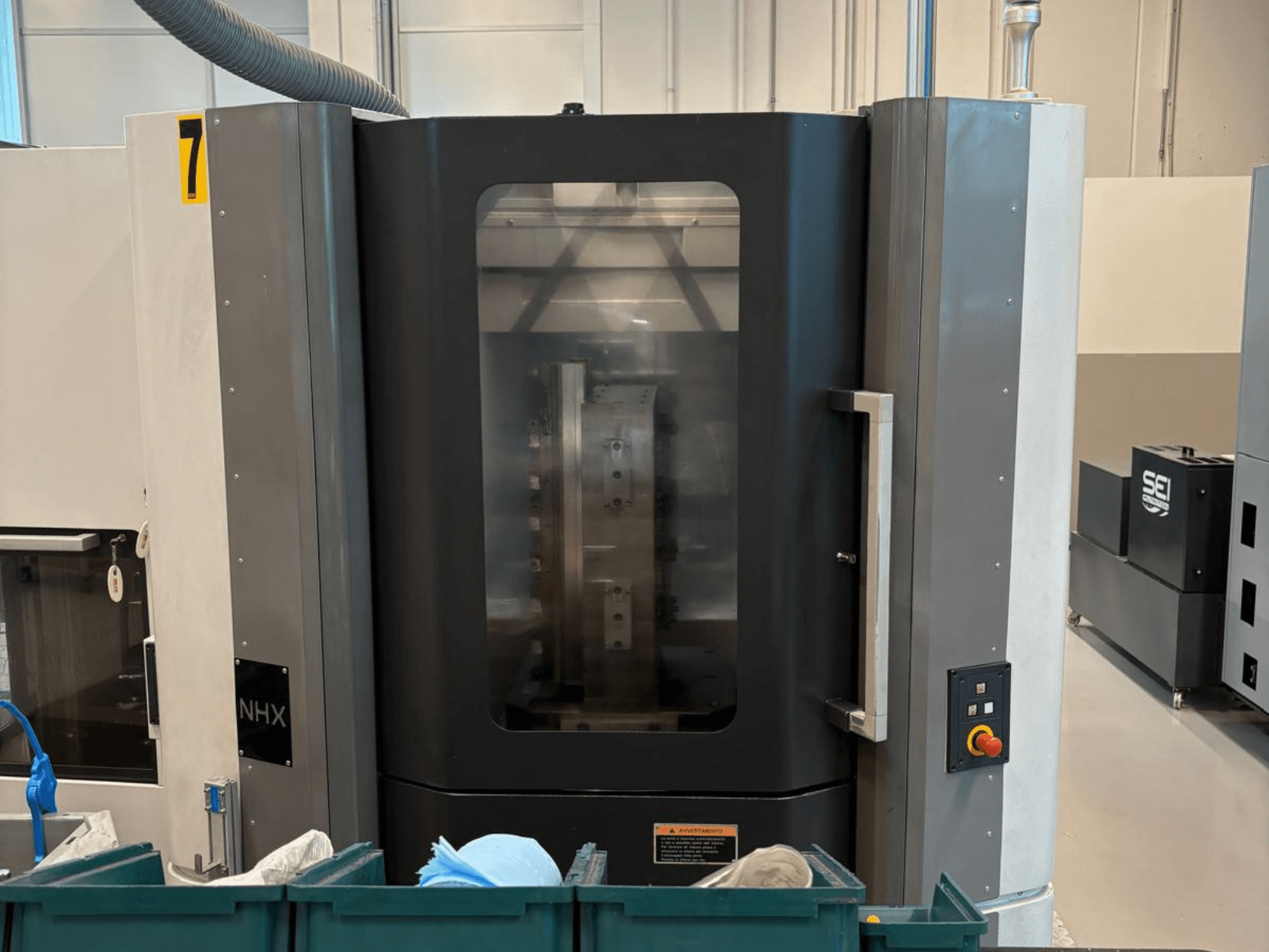 NHX CNC-gép elölnézetben, a vezérlőpanel és a jól látható kijelző; egy műhelyben található, zöld tárolókkal a közelben.