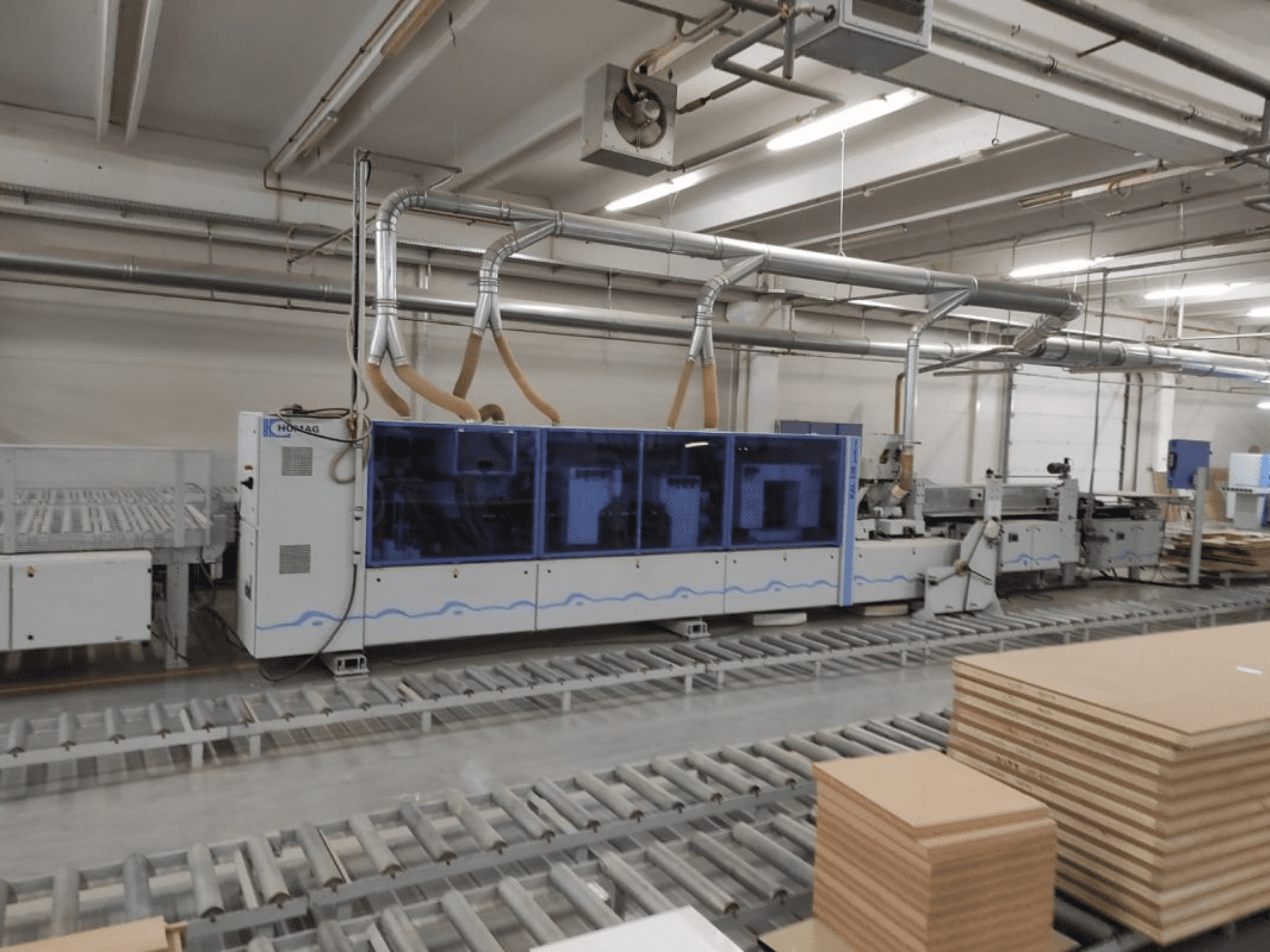 HOMAG gyártógép egy gyárban, elölnézetben, kék burkolattal és szállítószalagrendszerrel, egymásra helyezett rétegelt lemezekkel.