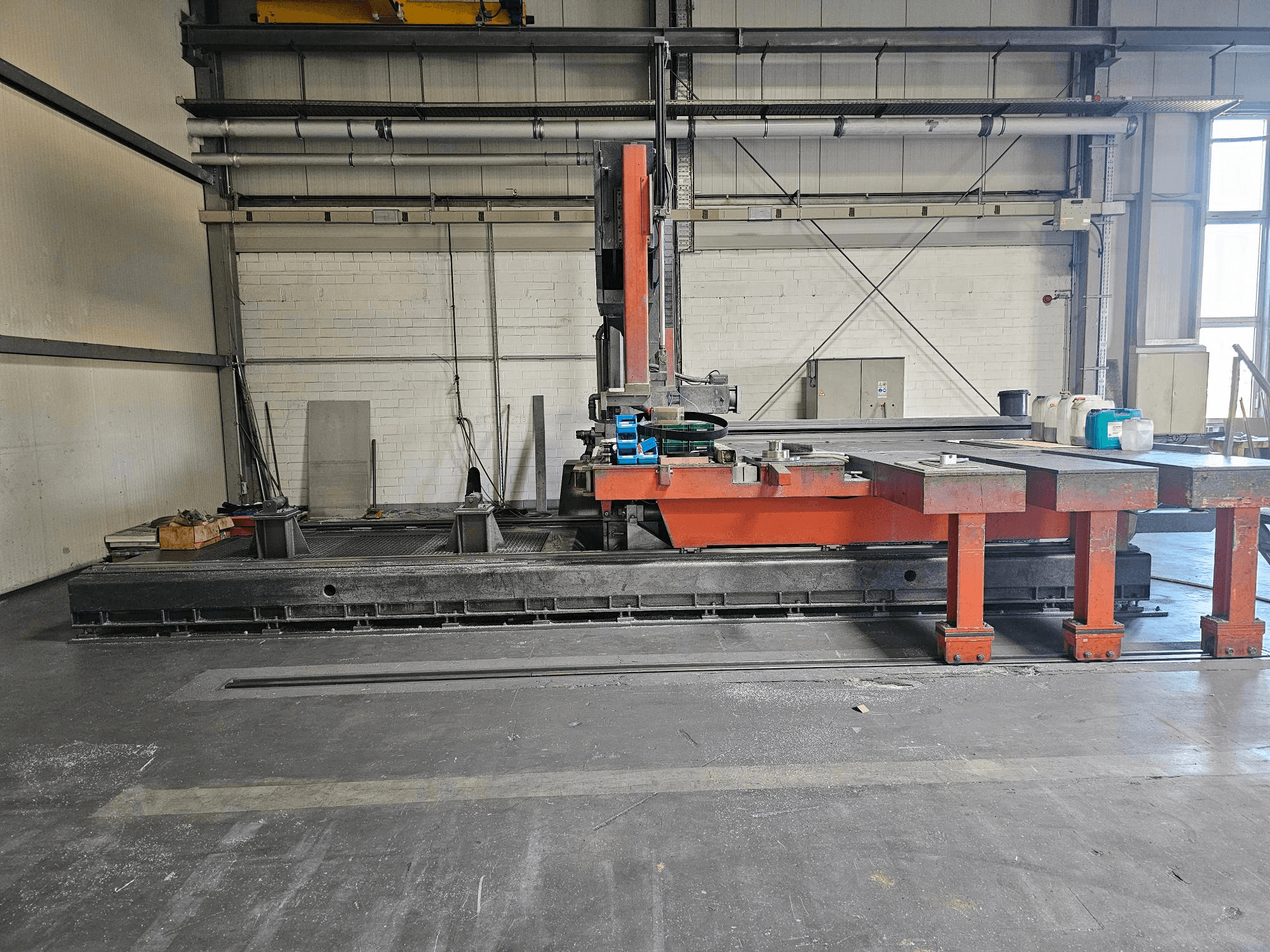 Ipari vágógép egy nyitott műhelyben, narancssárga és fekete színben, nagy asztallal; oldalnézetben a tartozékok és a munkaterület látható.