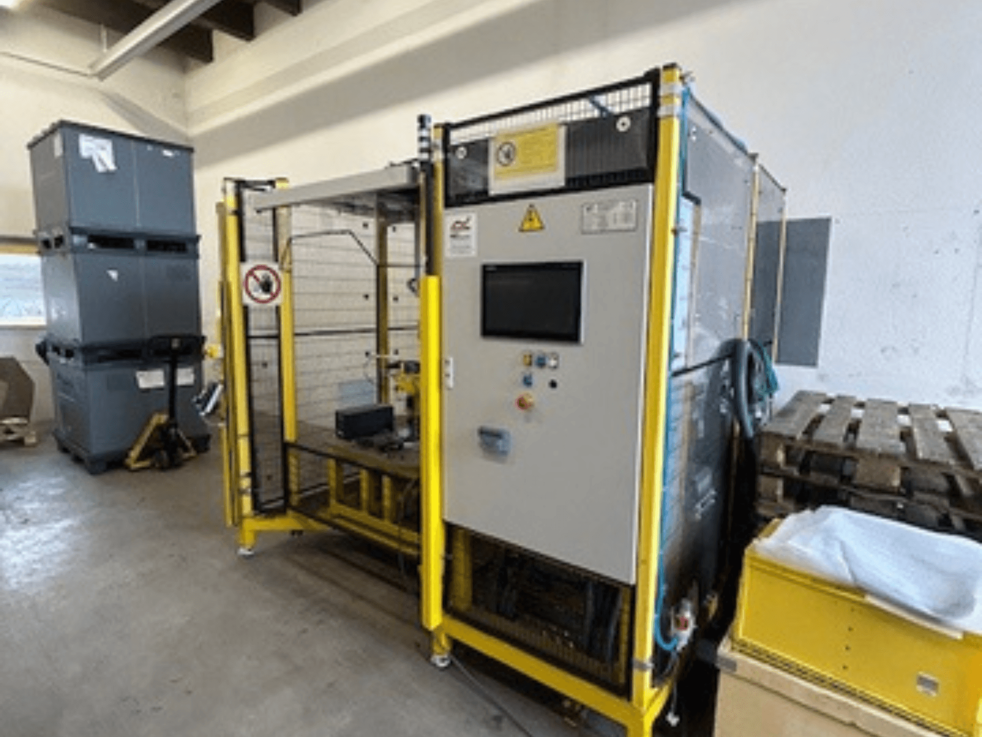 Ipari gép vezérlőpanellel és biztonsági berendezésekkel, elölnézetben; sárga keret, szürke tárolók és raklapok a háttérben.