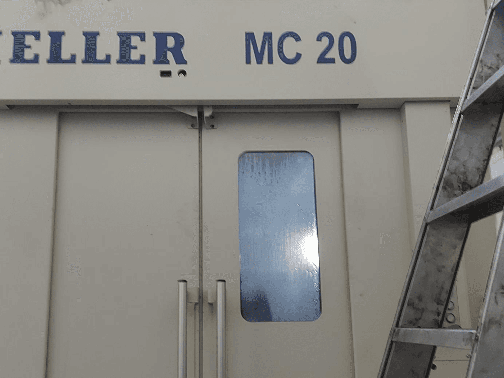 A HELLER MC 20 vízszintes megmunkálóközpont előlapja, amelyen az ajtókilincsek és egy kis ablak látható. Oldalán létra.