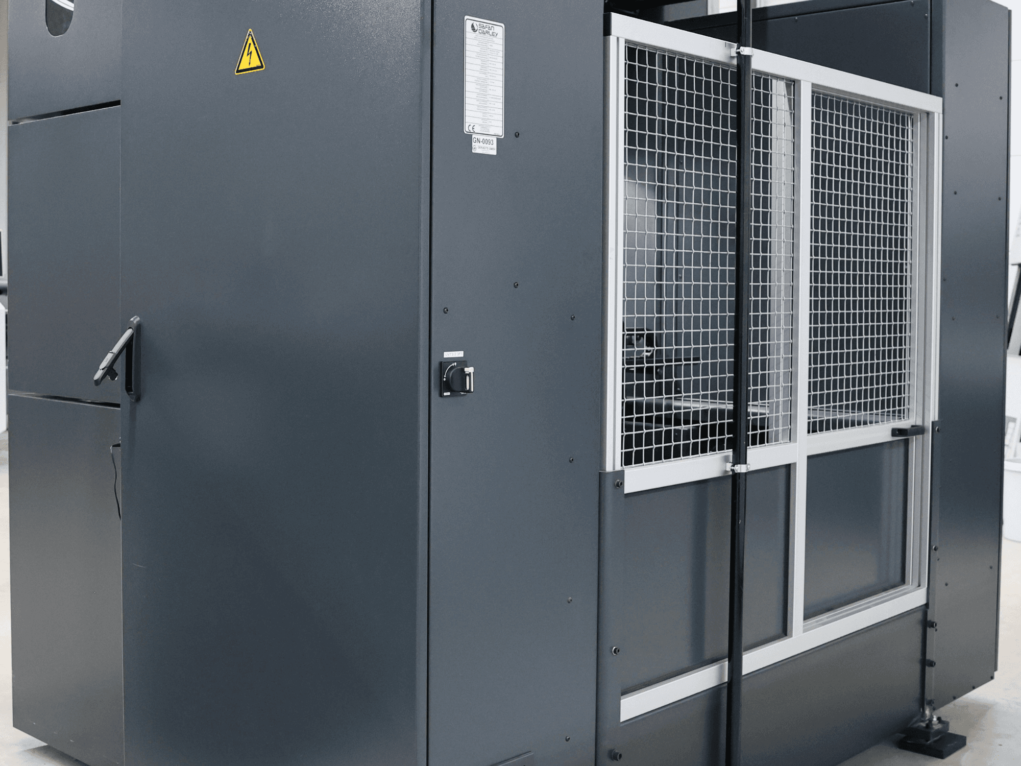 Szürke ipari gép biztonsági figyelmeztetéssel, elölnézetben a rácsos ajtó és a kezelőpanel látható, a modell fel van címkézve.