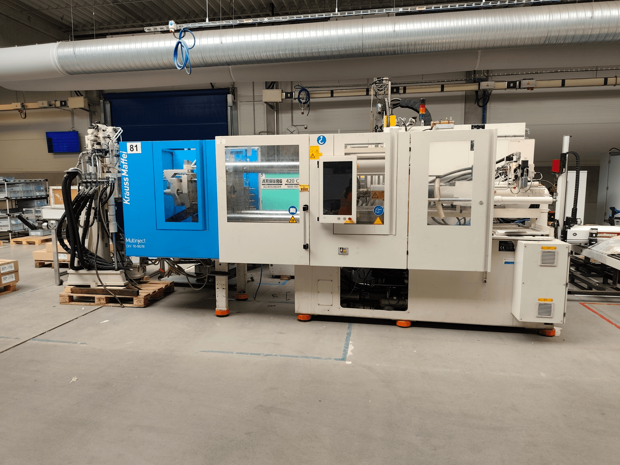 Krauss Maffei 110-180-180 CX V hidraulikus fröccsöntőgép, oldalnézet, amelyen látható a kék-fehér ház, a kezelőpanel és a csatlakozók.