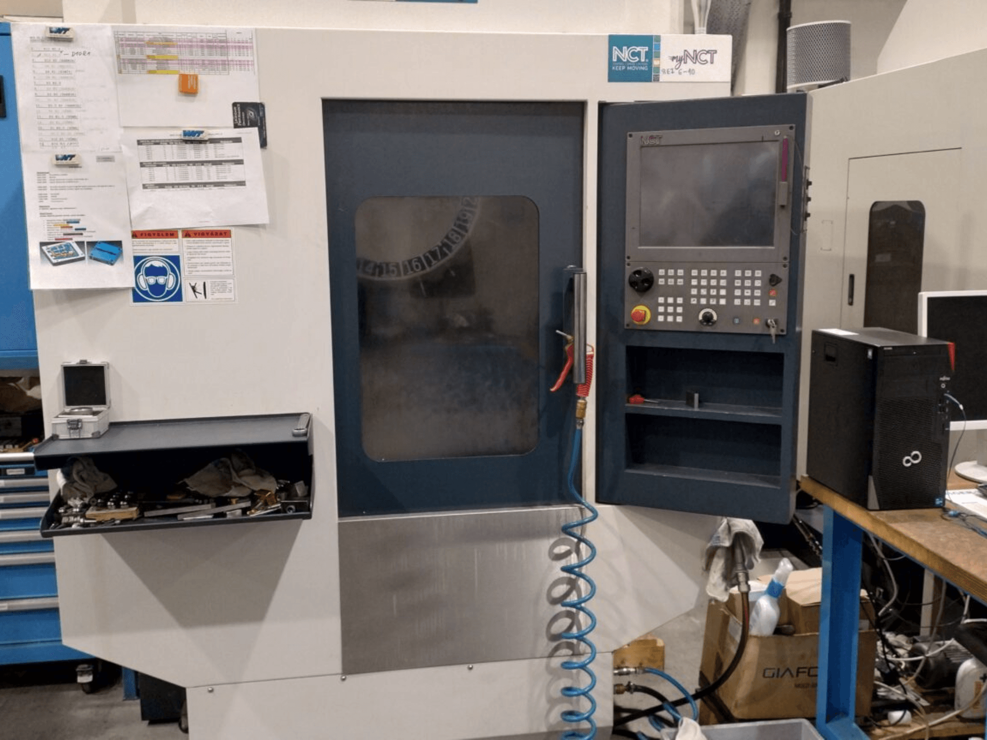 NCT CNC-gép, elölnézetben, vezérlőpanel numerikus billentyűzettel, szerszámok a tárolófiókban, kézikönyv a falon.