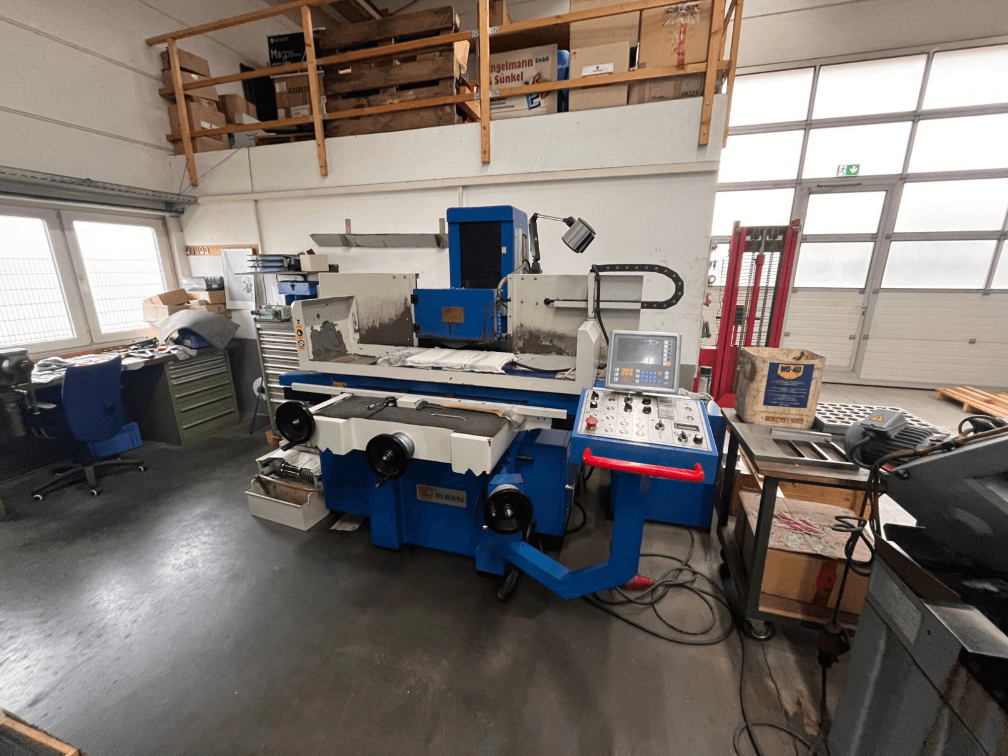 Kék felületcsiszoló gép digitális vezérlőpanellel, elölnézetben, a munkaterület rendetlenségével és egy zöld szerszámos szekrénnyel.