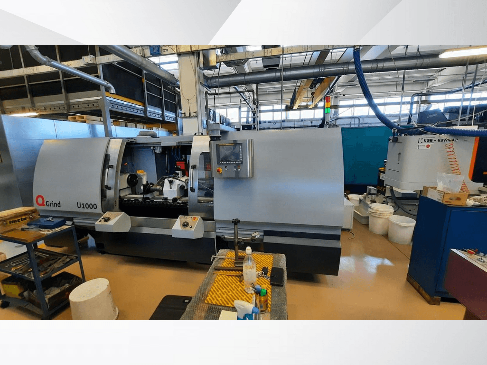 Q-Grind U1000 gép egy műhelyben, elölnézetben a vezérlőpanellel és a nyitott vágási területtel, körülvéve berendezésekkel.