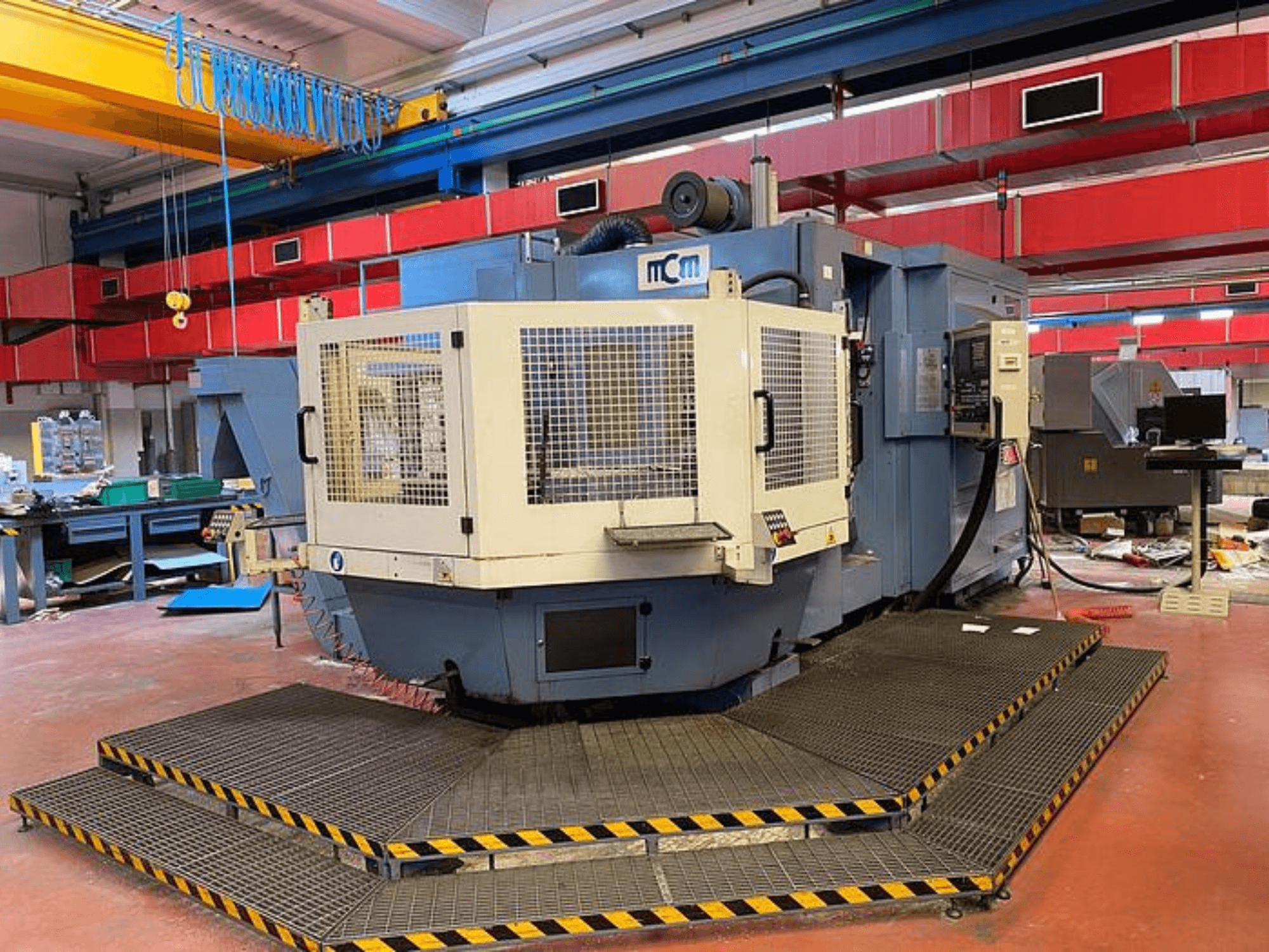 MCM CNC-gép egy gyárban, elölnézetben, védőketreccel, fekete és sárga biztonsági élekkel, ipari berendezésekkel körülvéve.