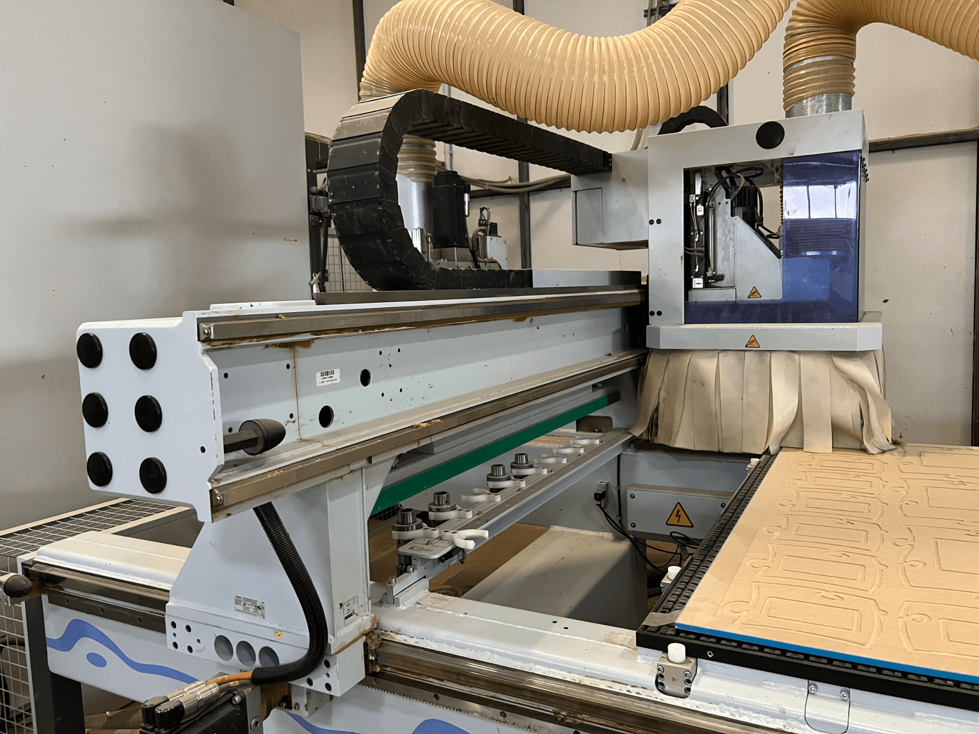 Egy kék és ezüst színű vezérlőpanellel ellátott, porelszívóval és fafaragó ágyazattal felszerelt CNC-router oldalnézetben.