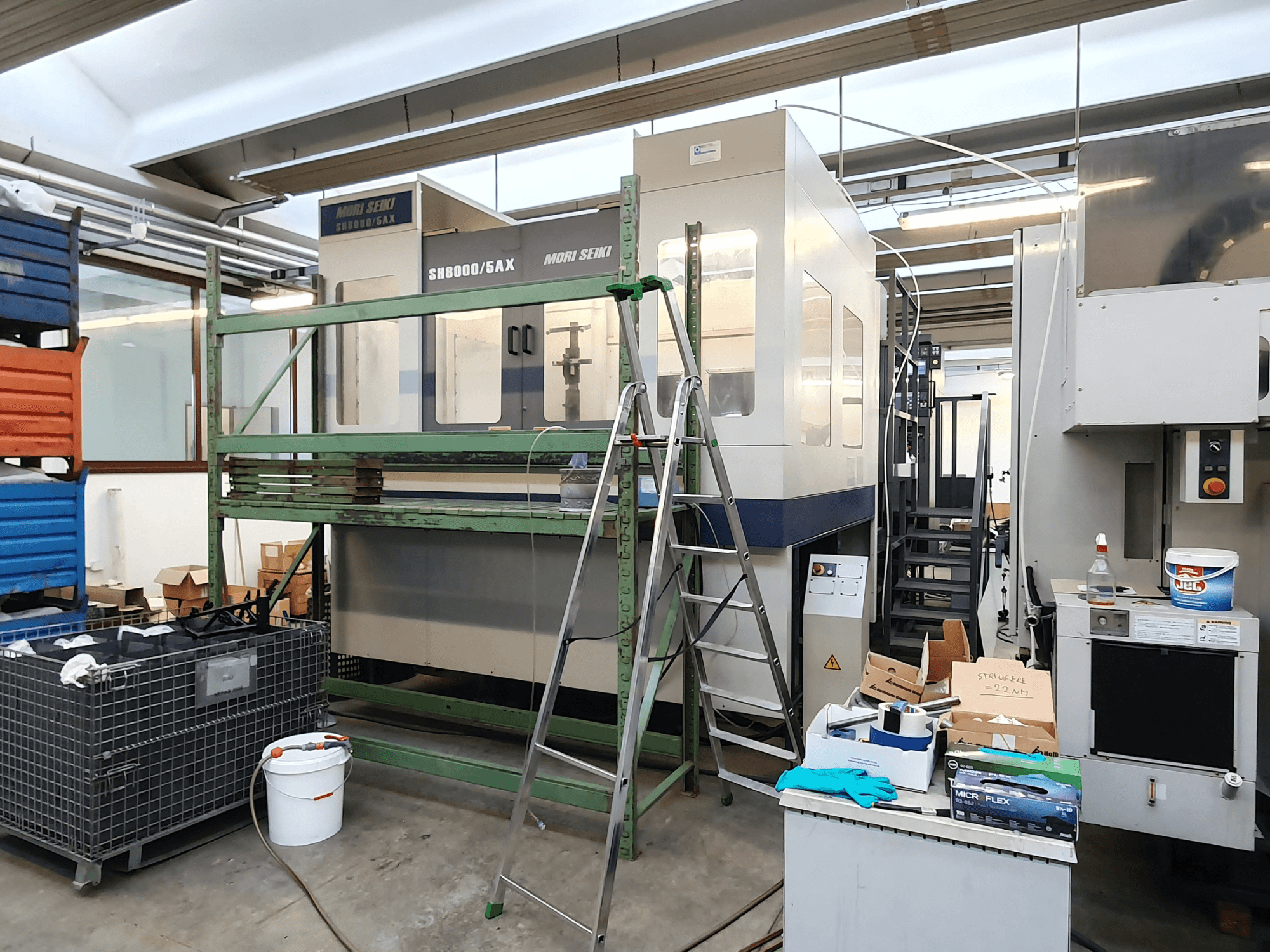 Mori Seiki SH-8000/5AX CNC-gép egy műhelyben, elölnézetben, műhelyszerszámokkal és tároló dobozokkal a közelben.