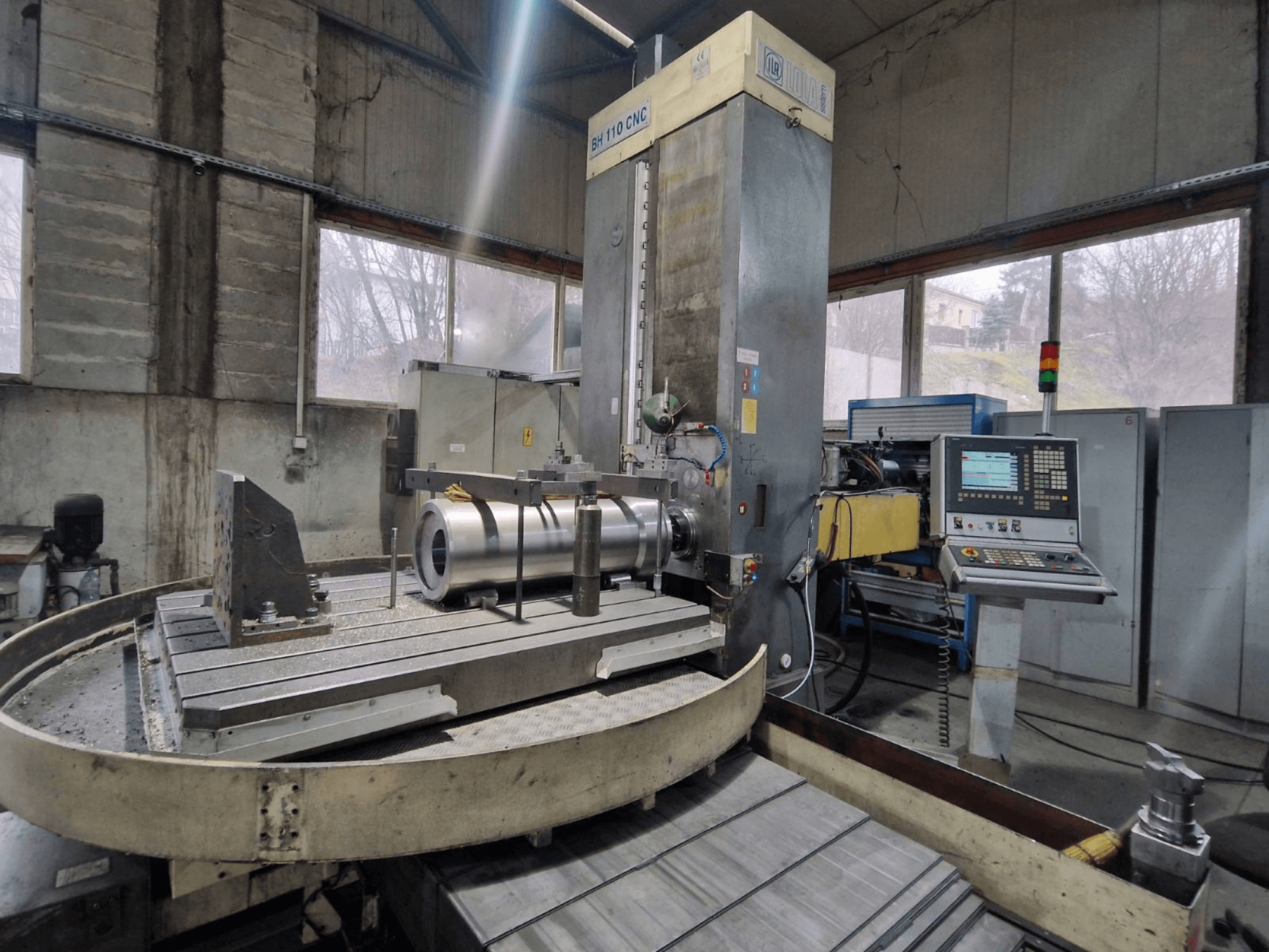 LOLA BH 110 CNC fúrógép egy műhelyben, elölnézetben, fém munkadarab és vezérlőpanel.