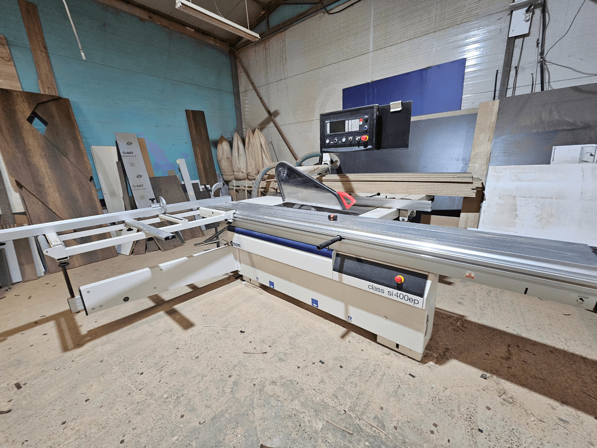 SI400EP osztályú lemezfűrész, elölnézetből, állítható kerítéssel, homokos műhelypadlóra állítva, falapokkal körülvéve.