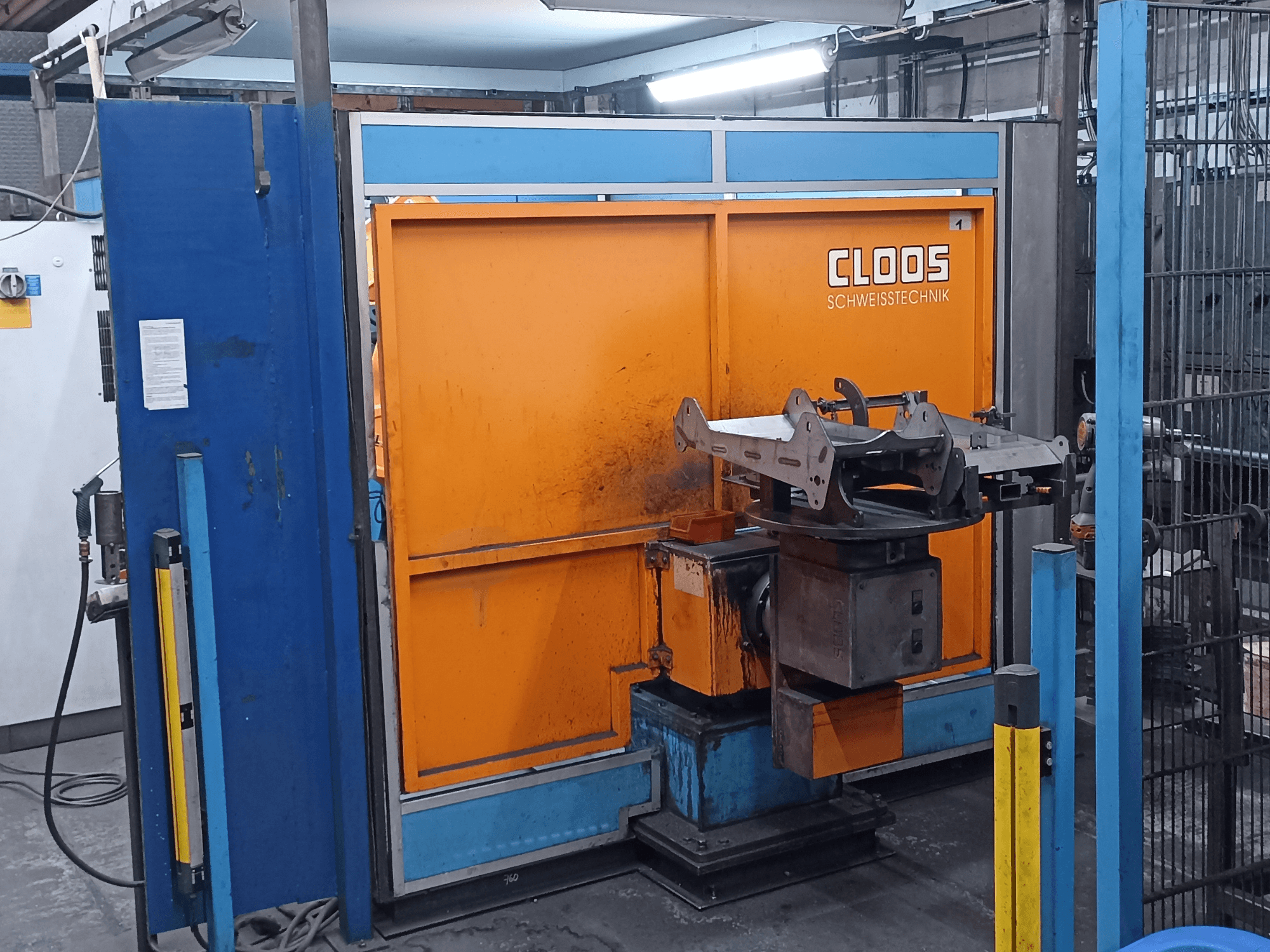 CLOOS Romat 320 hegesztőrobot egy műhelyben, elölnézetből, narancssárga vezérlőpanellel és egy forgó talapzaton elhelyezett fém munkadarabbal.