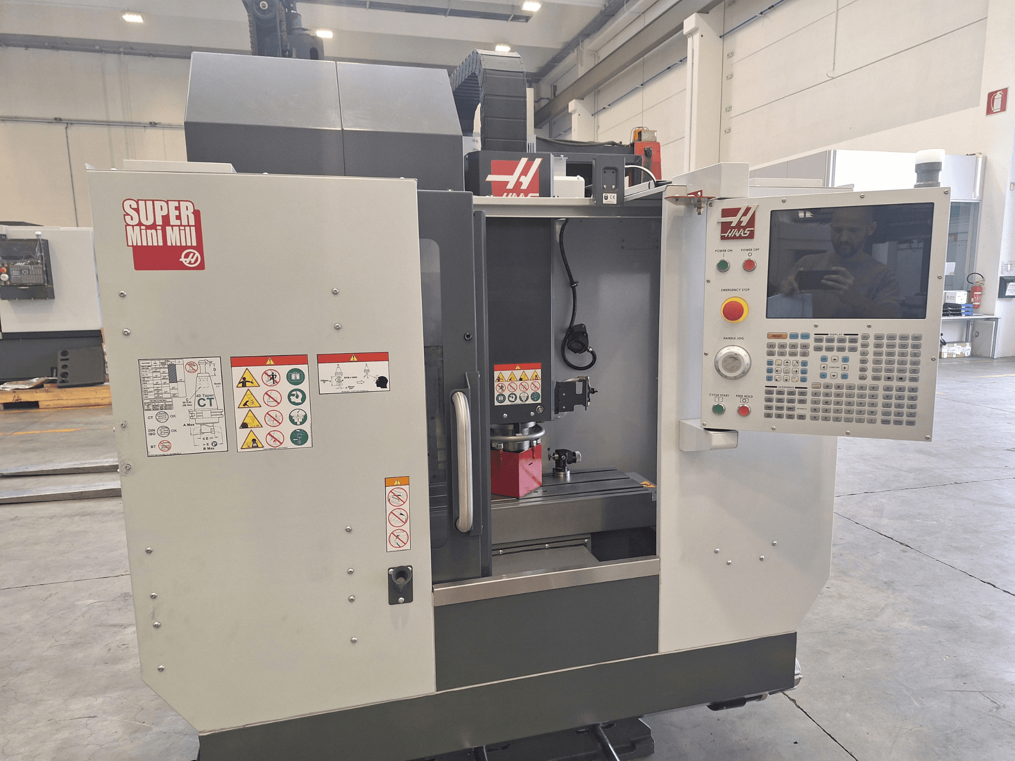 A HAAS Super Mini Mill elölnézete, amelyen látható a vezérlőpanel, a biztonsági címkék és a megmunkálási terület egy beállított munkadarabbal.