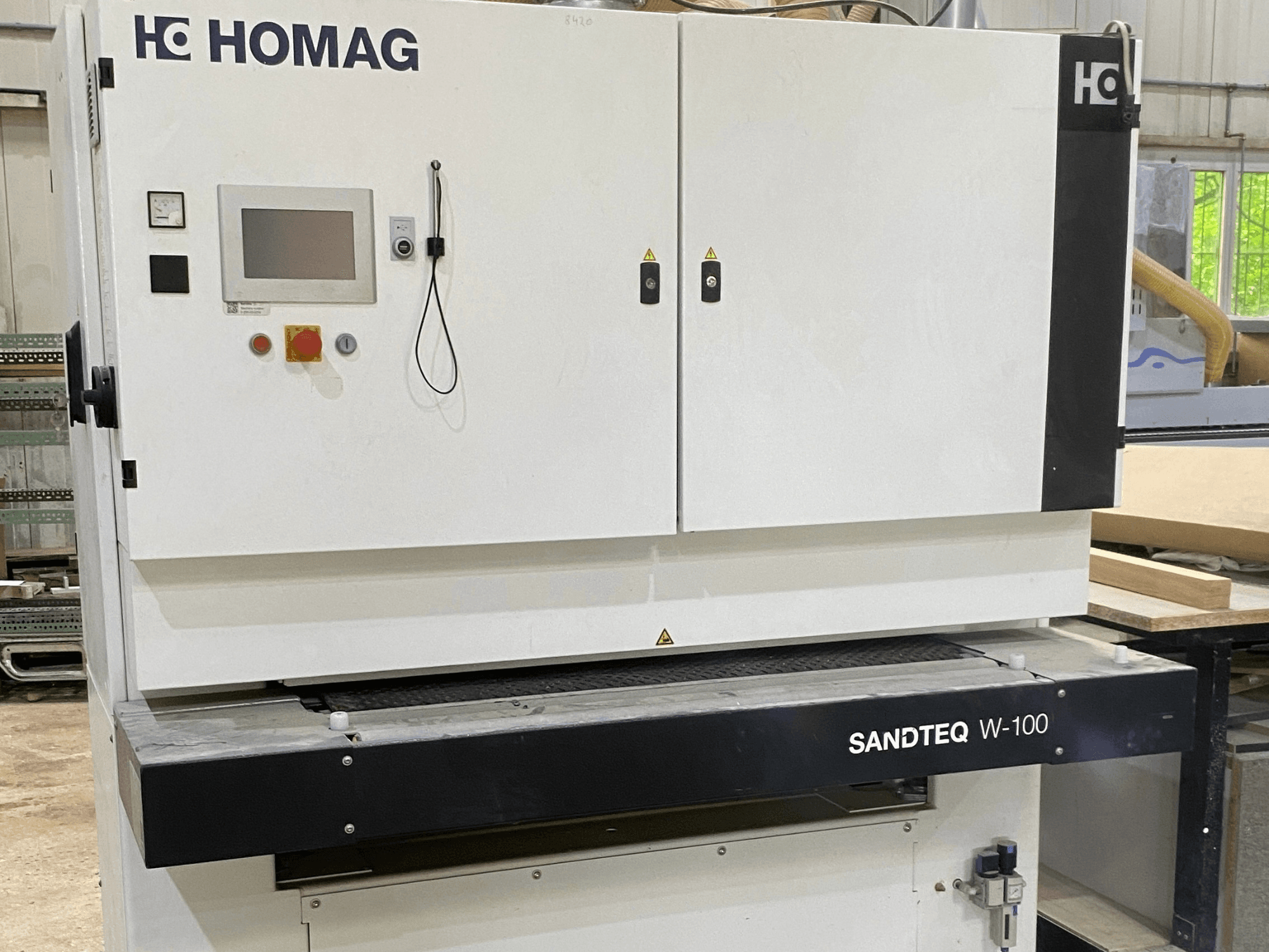 HOMAG SANDTEQ W-100 szalagcsiszoló egy műhelyben, látható a vezérlőpanel, a porelszívó csatlakozó és a csiszolószalag területe. Elölnézet.