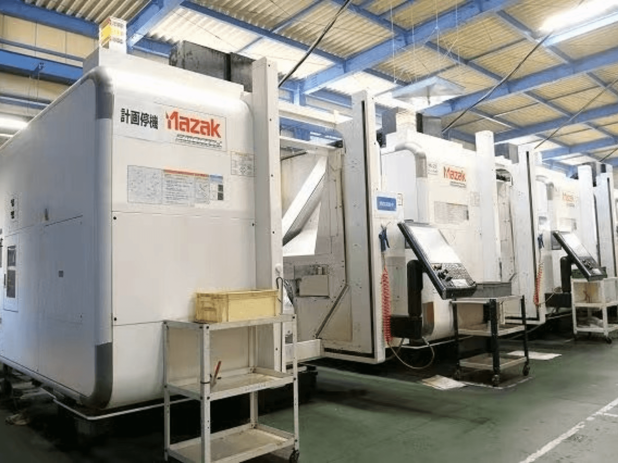 Mazak CNC-gépek gyári környezetben, oldalnézetben a vezérlőpanelek és címkék fehér testeken.