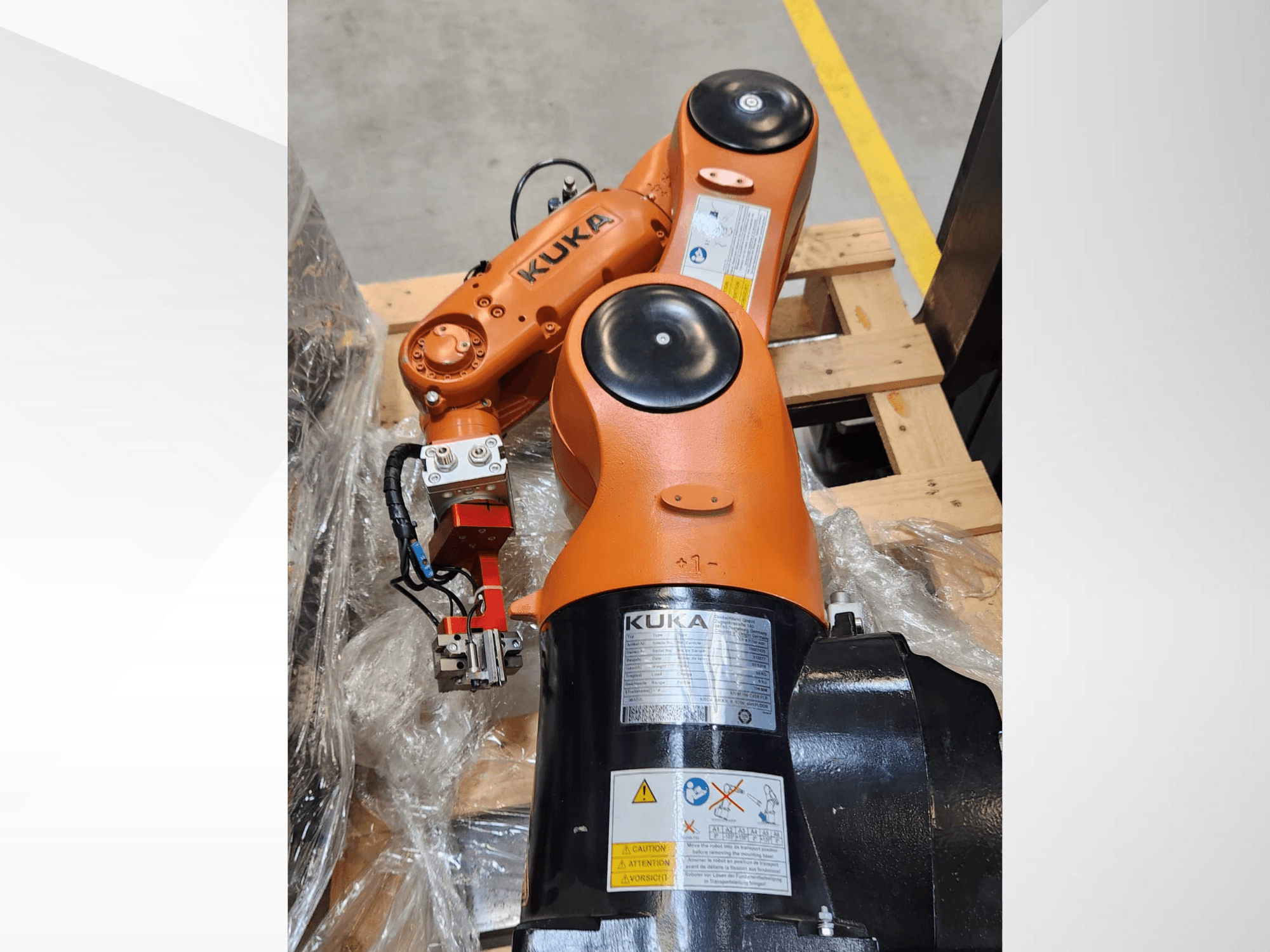 KUKA robotkar narancssárga kivitelben, oldalnézetben, megfogóval és vezérlő vezetékekkel egy fa raklapon.