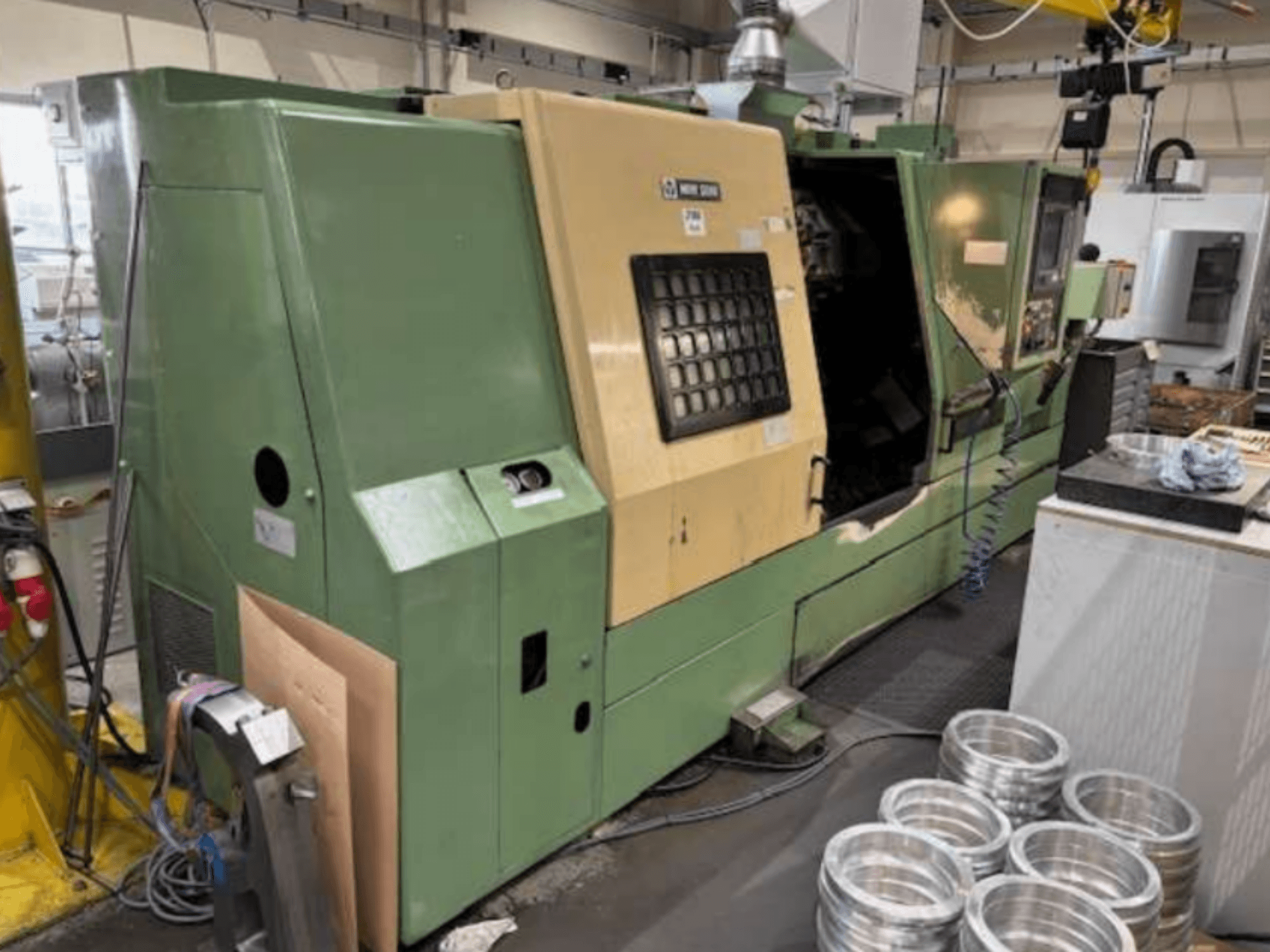 A Mazak zöld CNC esztergagépe szemből nézve, a vezérlőpanel és a megmunkált alumínium alkatrészek a munkaasztalon.