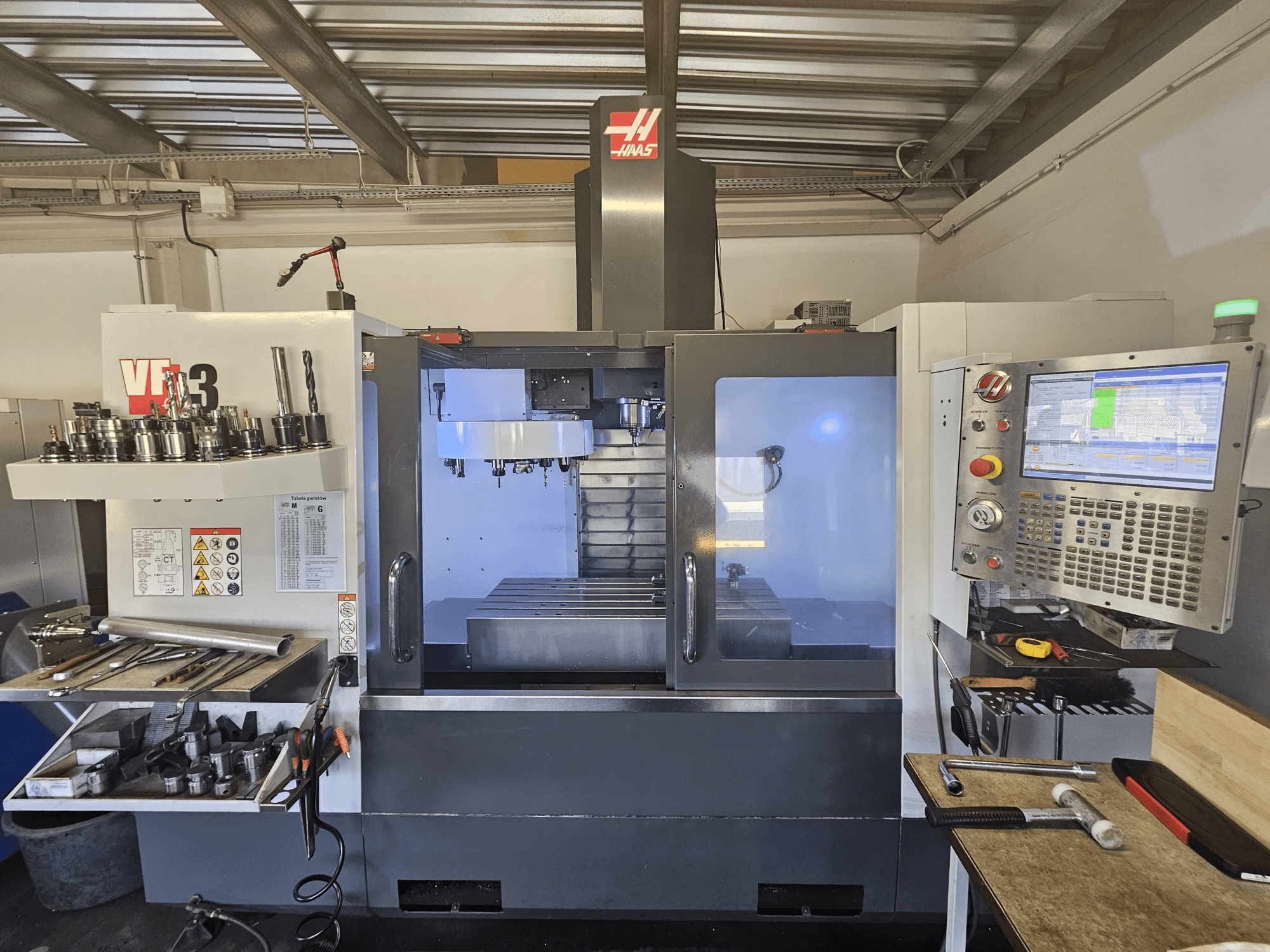Haas VF-3 CNC-gép egy műhelyben, elölnézetben a szerszámok, a vezérlők és a marási terület látható, megvilágított belső térrel.