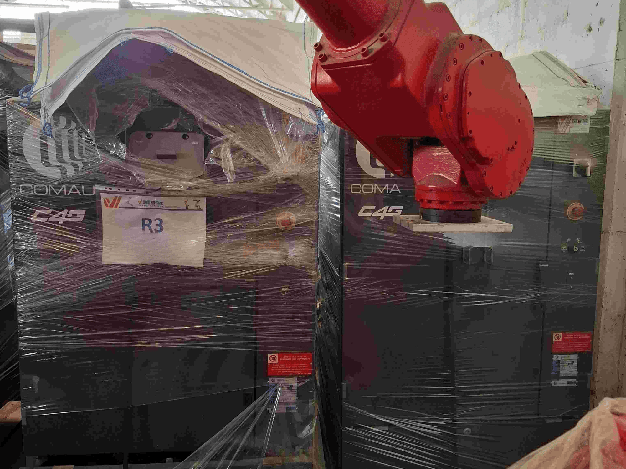 Comau C4G robotkar szemből nézve, műanyagba csomagolva, "R3" felirattal, a gépen látható figyelmeztető matricákkal.