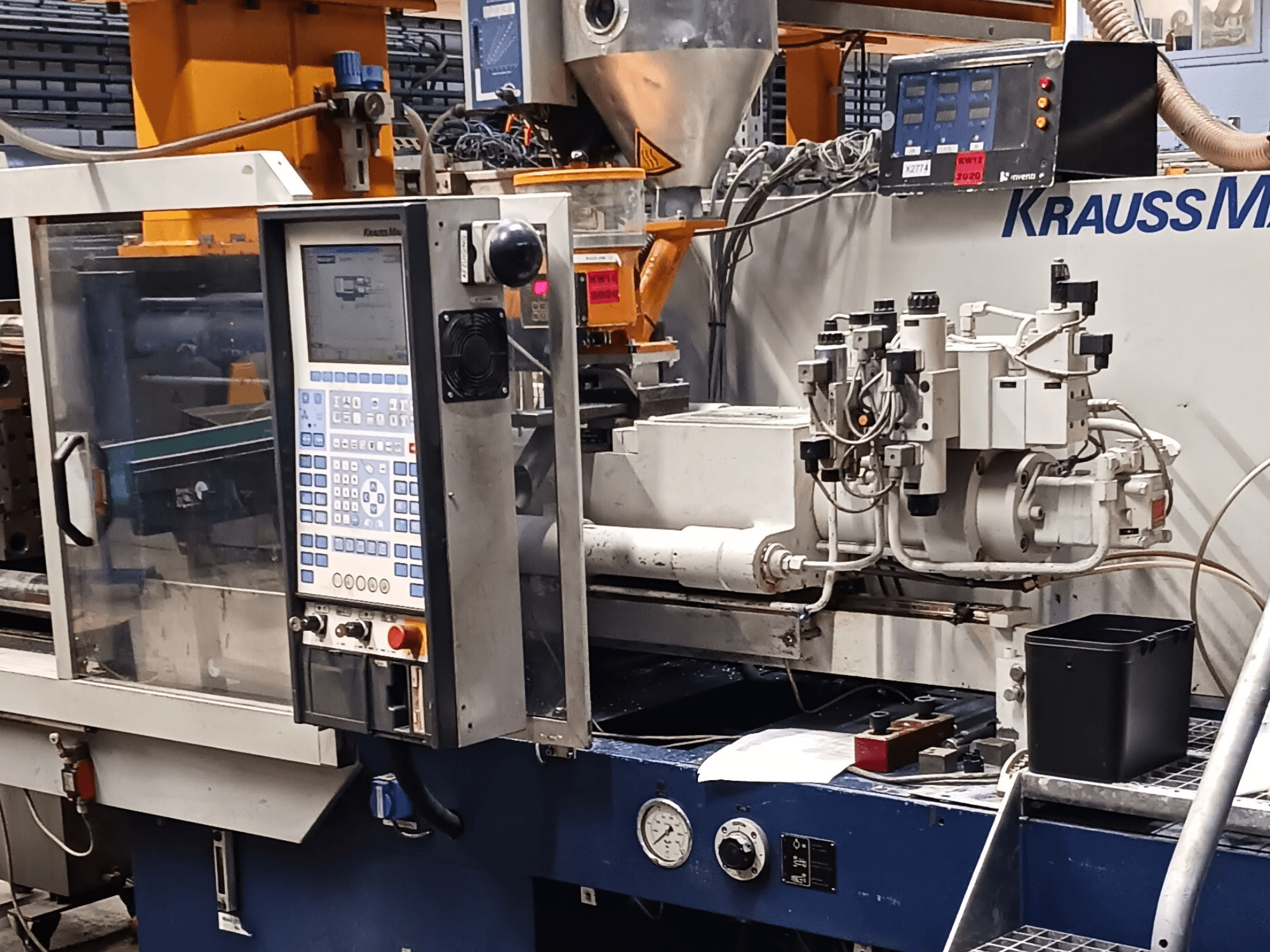 KraussMaffei fröccsöntőgép, előtérben a vezérlőpult, a hidraulikus egységek és a szerszámok, elölnézetben.