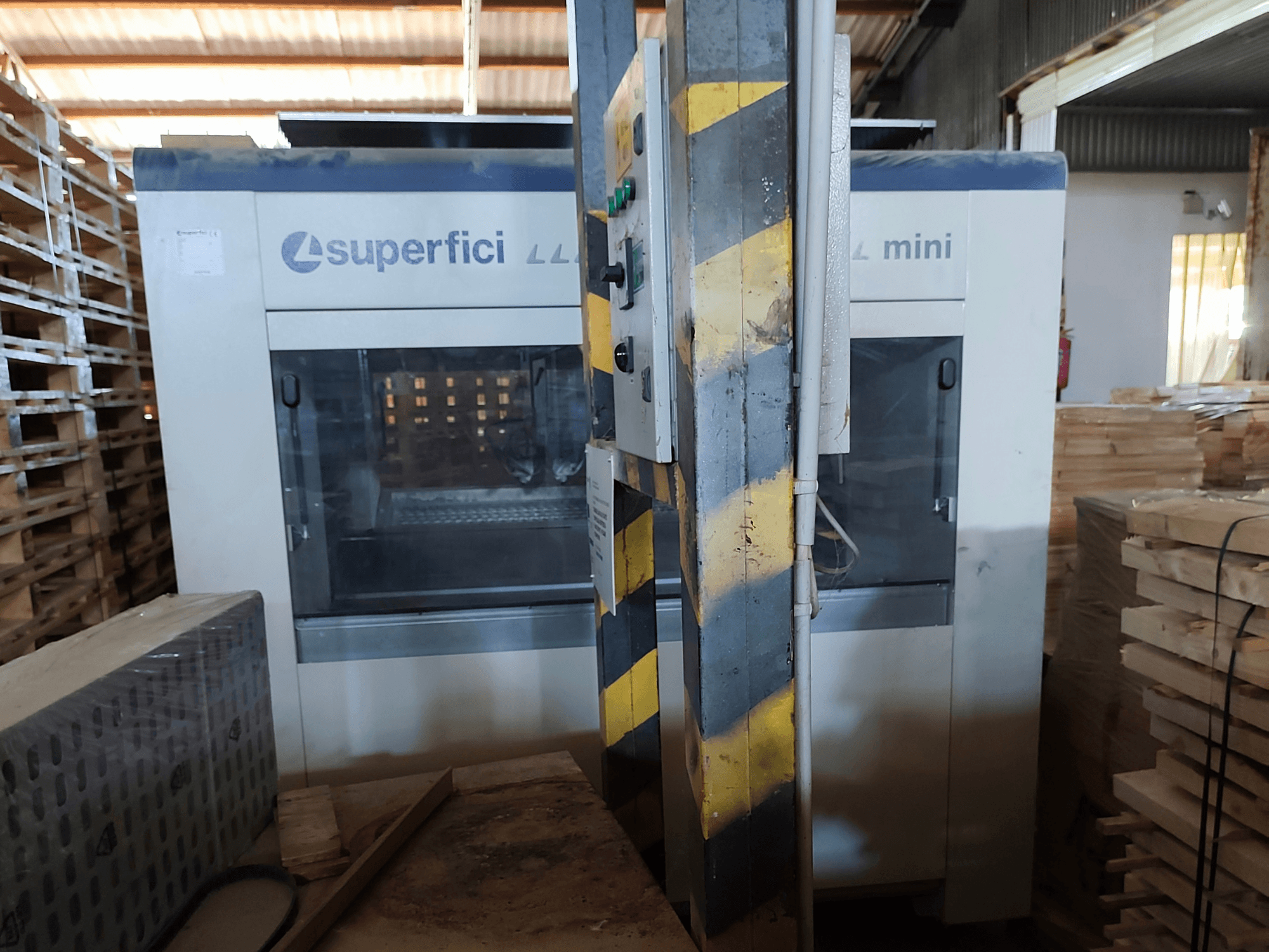 Az SCM Superfici Mini faipari felületkezelő gép elölnézetben, a vezérlőpanellel és a körülötte elhelyezett fa raklapokkal.