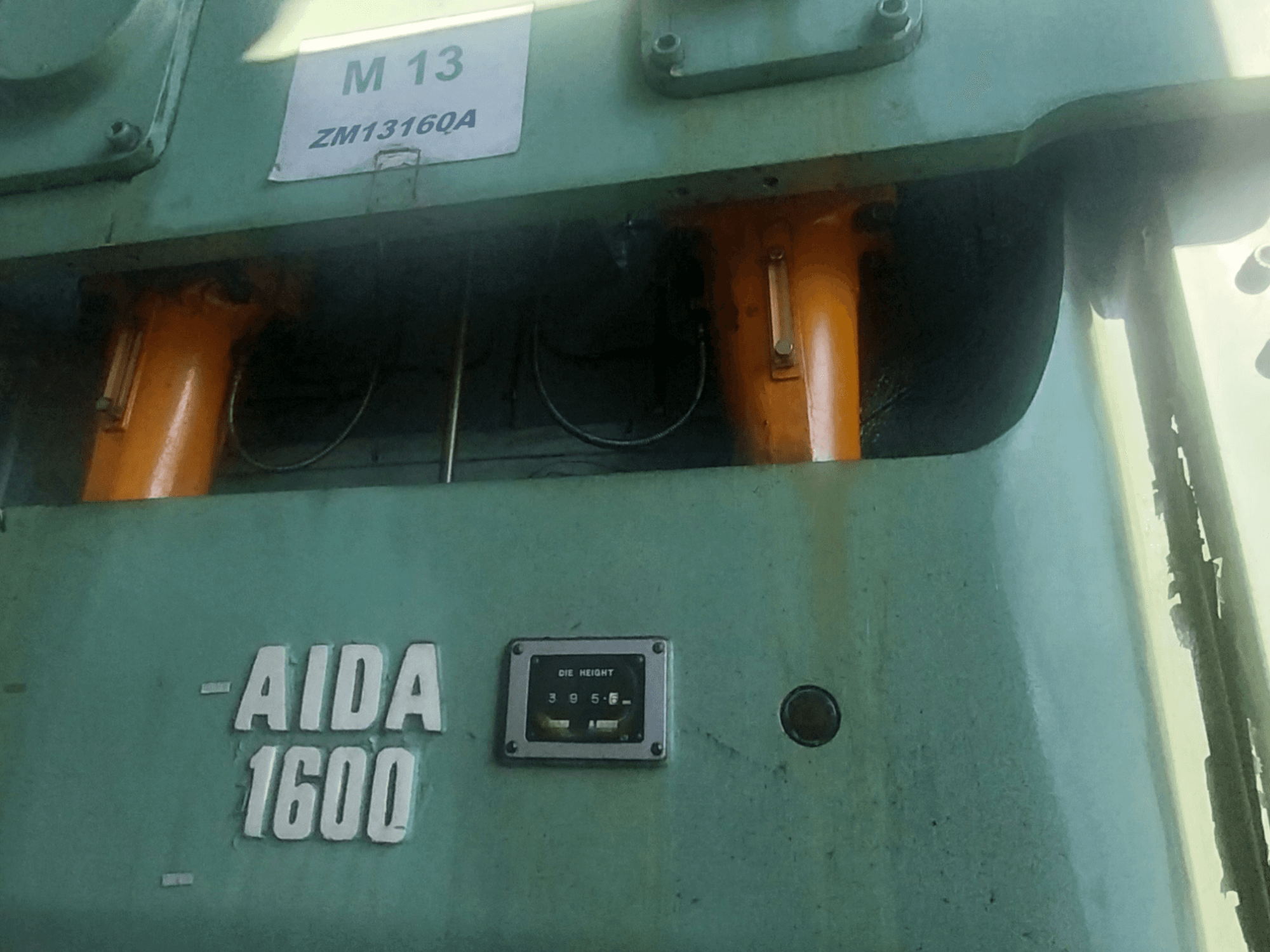 AIDA NC2-1600 mechanikus bélyegzőprés, elölnézet; láthatóak a narancssárga hidraulikus alkatrészek és a szerszámmagasságmérő.
