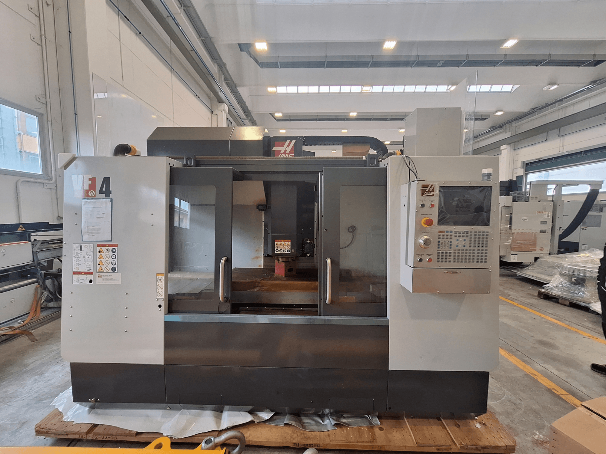 HAAS VF-4 függőleges megmunkáló központ, elölnézet, amelyen látható a vezérlőpanel, a munkaterület és a biztonsági címkék egy raktári környezetben.