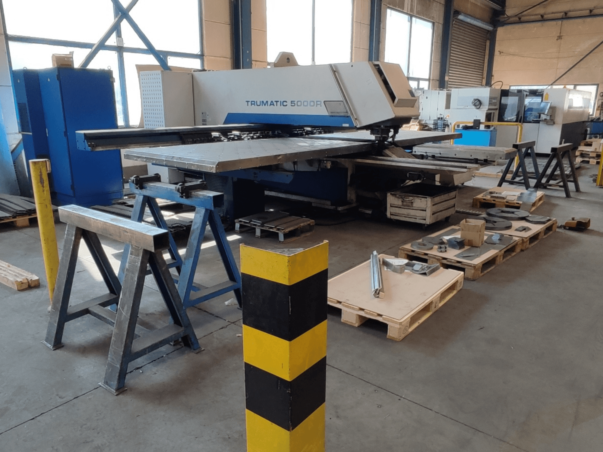 TRUMPF Trumatic 5000 R CNC-lyukasztógép egy műhelyben, oldalnézetből, a közelben raklapokon elhelyezett szerszámokkal és anyagokkal.