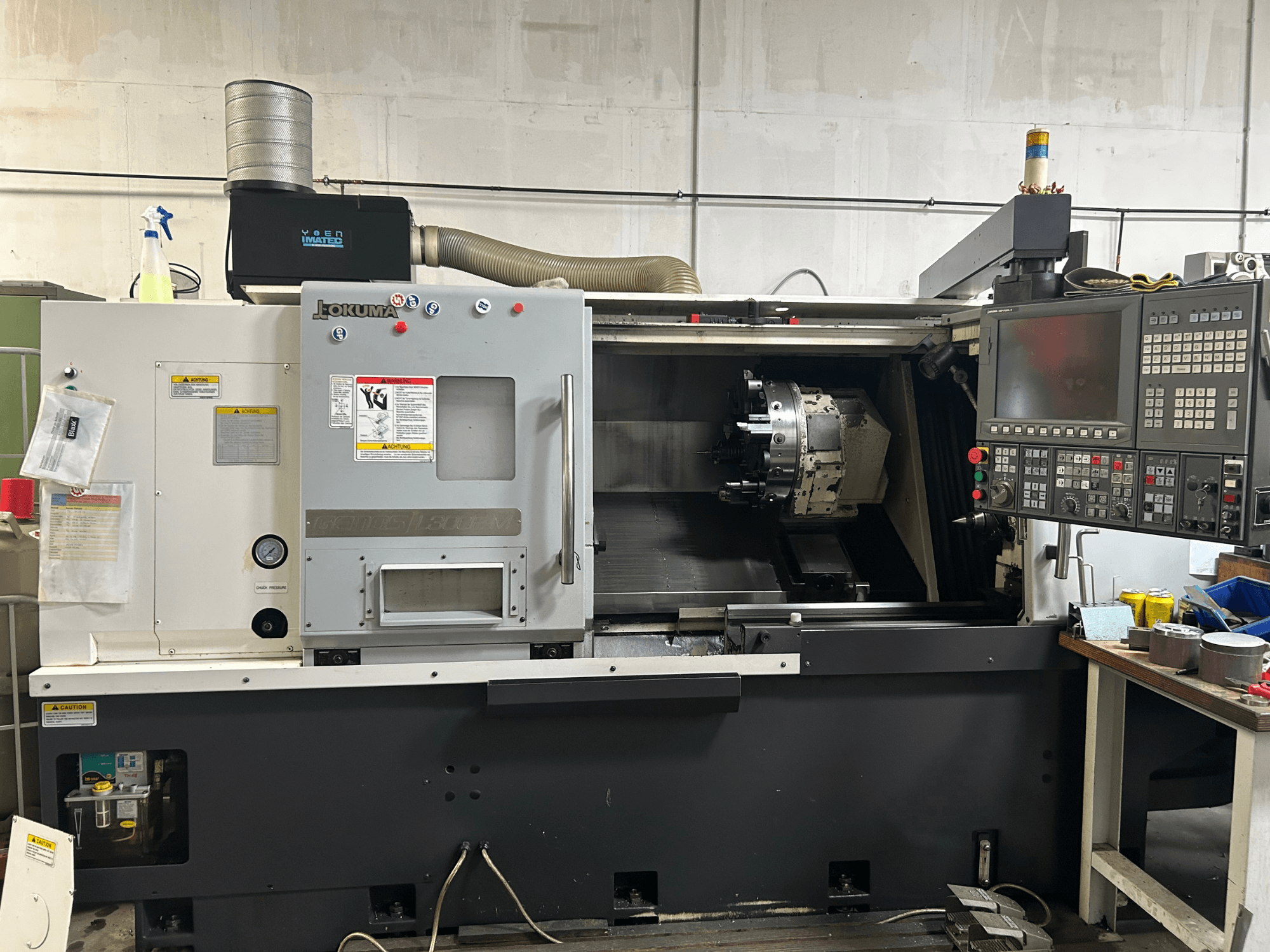 Egy Okuma Genos L300E-M vízszintes esztergagép elülső nézete vezérlőpanellel, látható orsóval és különböző beállítási szerszámokkal.