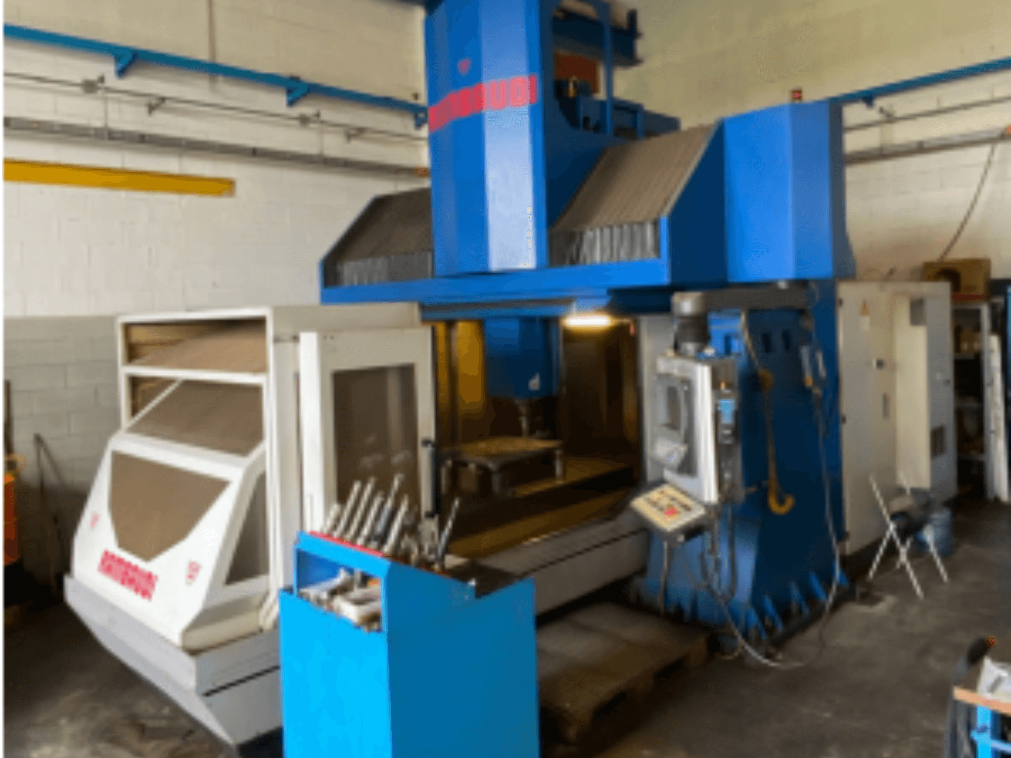 Függőleges megmunkálóközpont Rambaudi RF103, elölnézetben a kezelőszervek, a szerszámtároló és a megmunkálási terület. Kék és fehér külső.