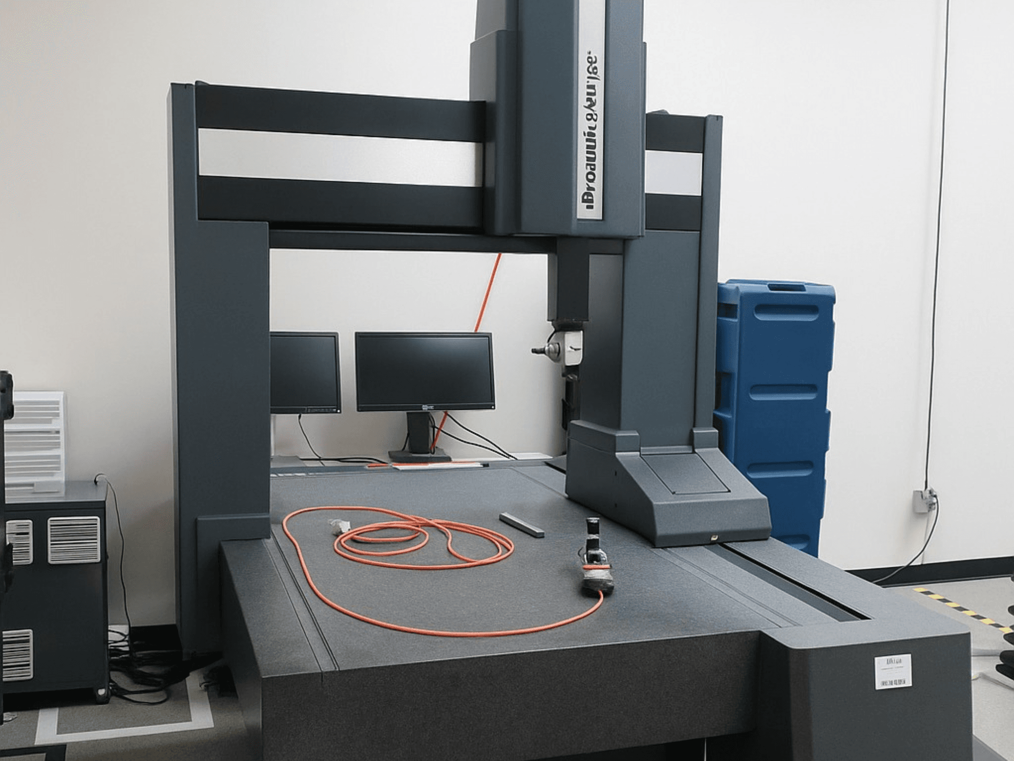 Brooks Automation koordináta-mérőgép két monitorral, elölnézetben, szürke asztallappal és mérőeszközökkel.