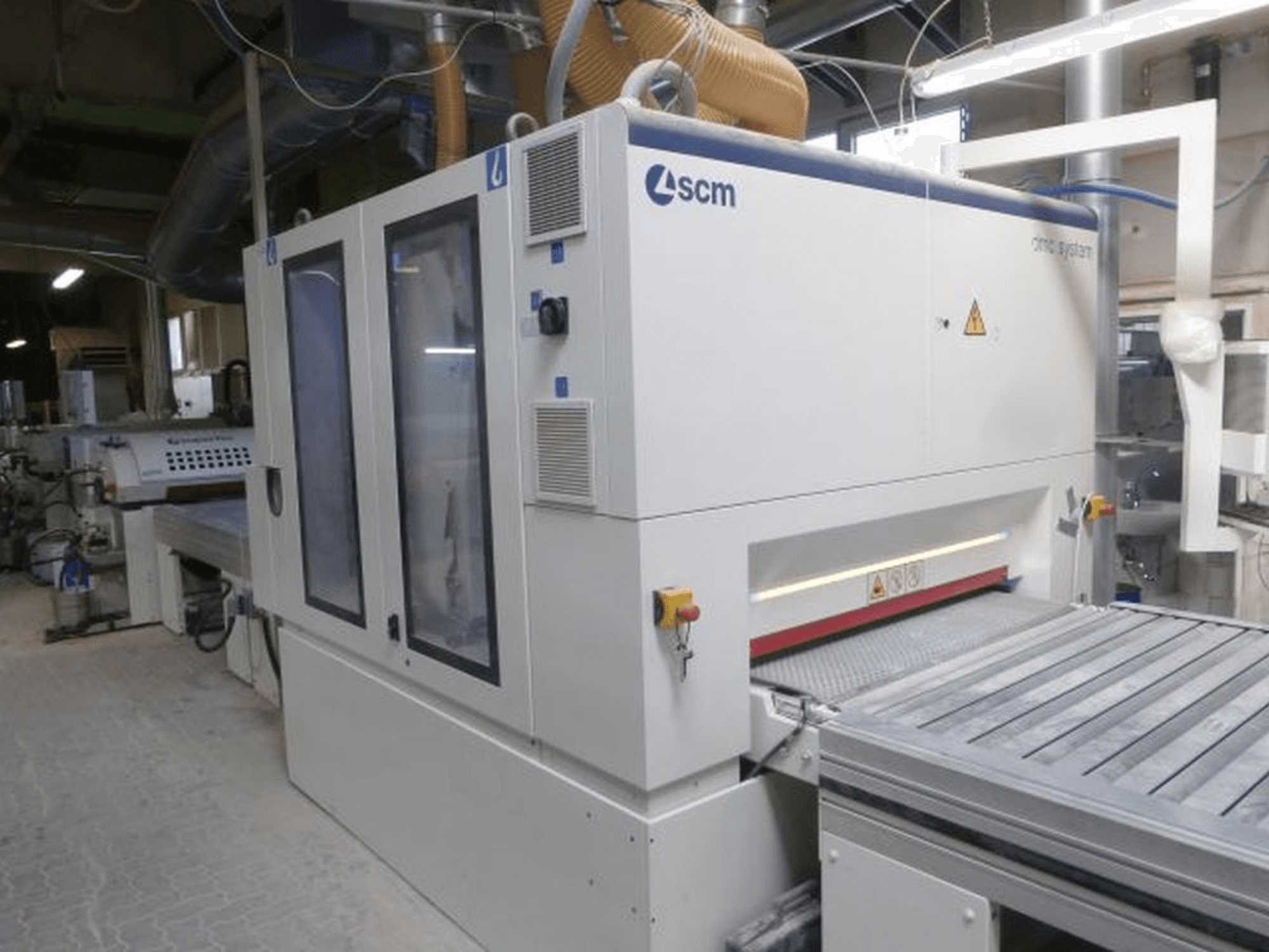 SCM DMC SY TC 1350 szalagcsiszoló egy műhelyben, oldalnézetből, amelyen látható a vezérlőpanel és a csiszolófelület a biztonsági berendezésekkel együtt.